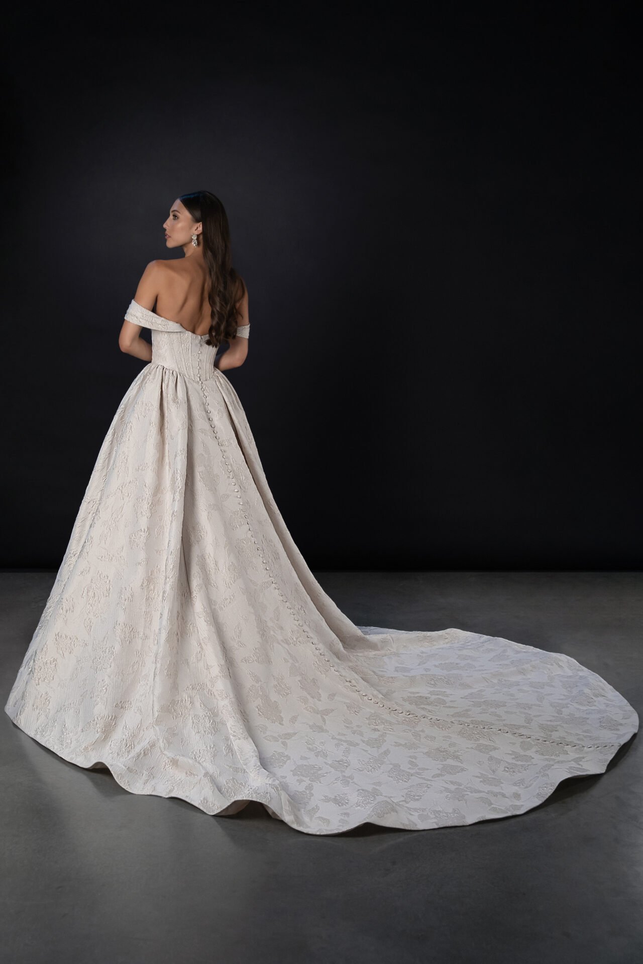 A woman in an off-the-shoulder ivory wedding gown with a long train, standing against a dark background, facing sideways with her hair down.