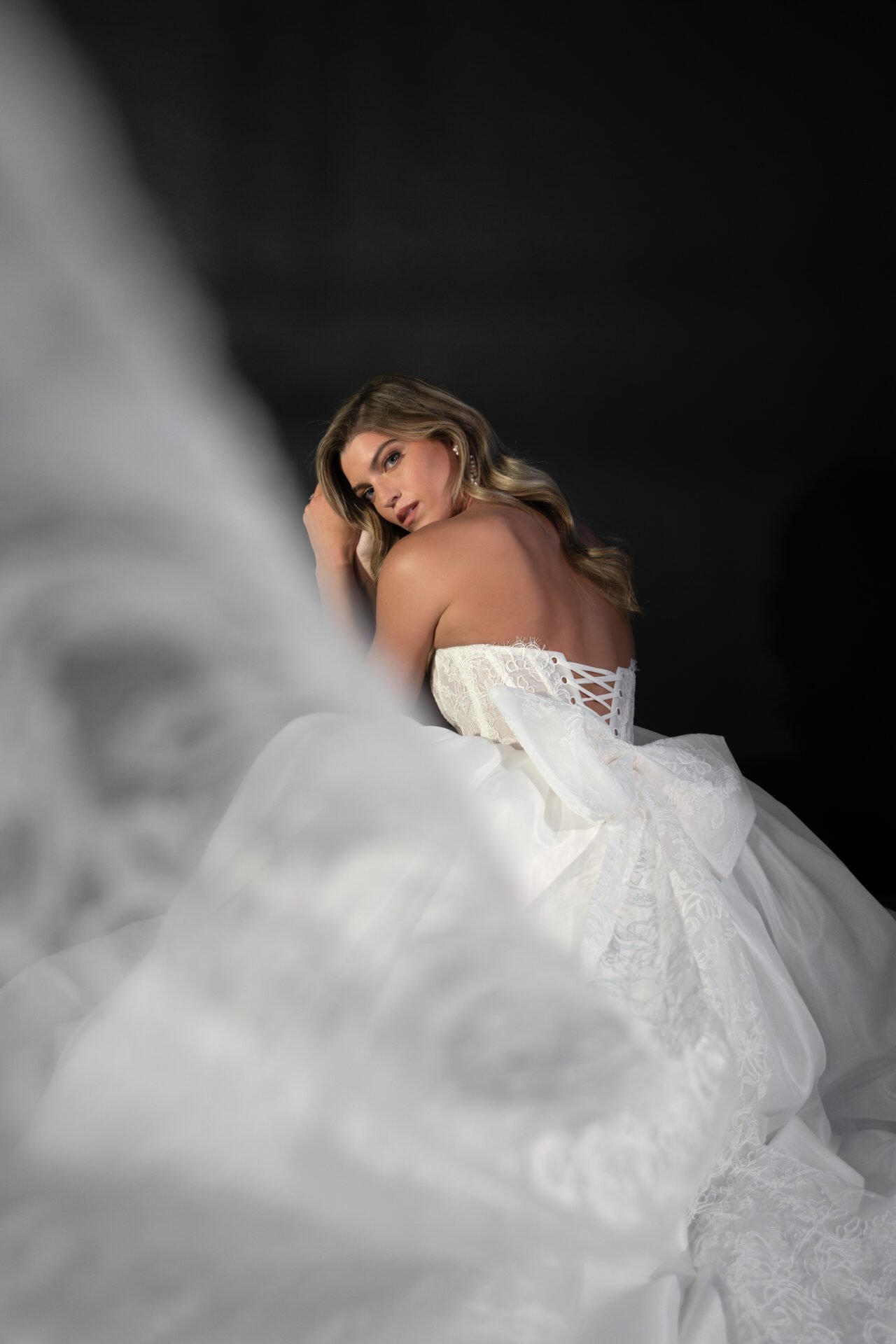A woman in a white wedding dress with lace-up back detail, sitting with her head resting on her arm, against a dark background.