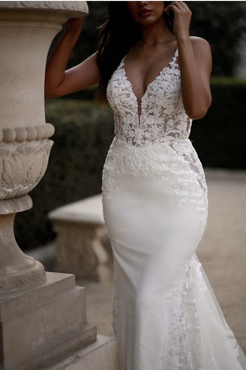A woman in a fitted, white wedding dress with lace detailing and a deep V neckline, standing outdoors near stone columns and benches.