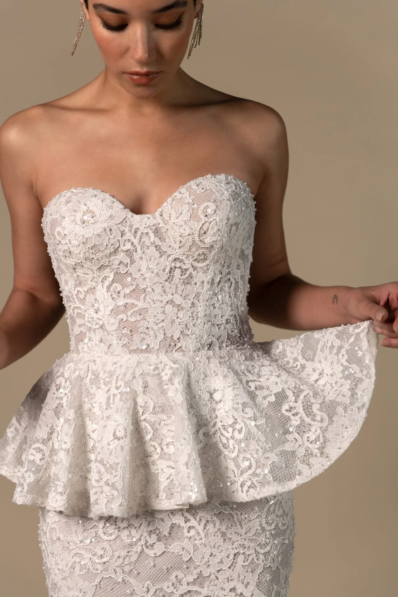 A woman wearing a strapless, lace, cream-colored dress with intricate floral embroidery and sequins, holding the bottom of the dress's layered peplum detail.
