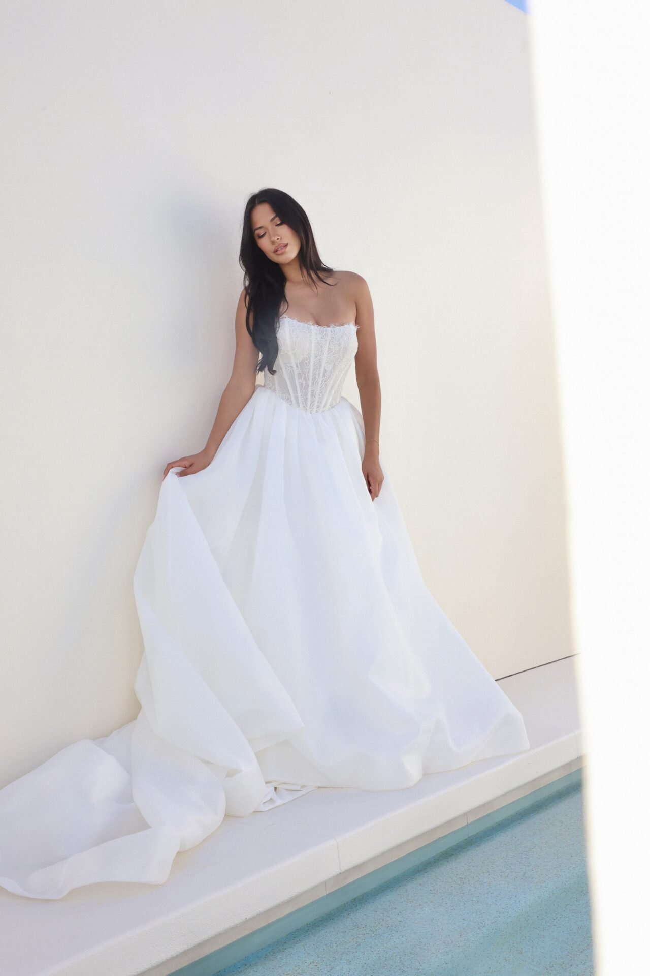 A woman in a white wedding dress standing against a plain white wall, with her eyes closed and one hand lifting the dress slightly.