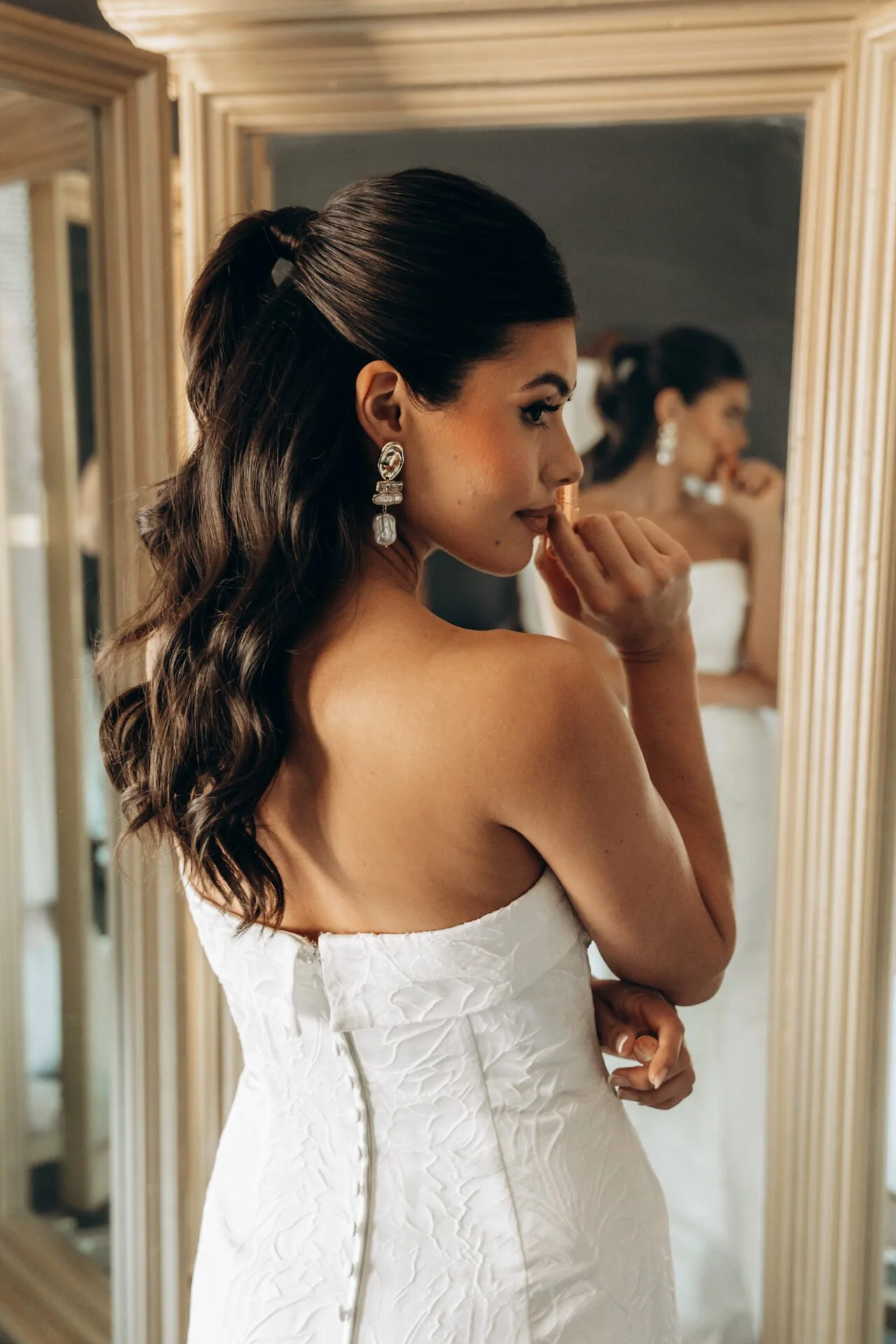 A woman in a white strapless wedding dress with dark wavy hair and large earrings, standing in front of a mirror, touching her lips with one hand.