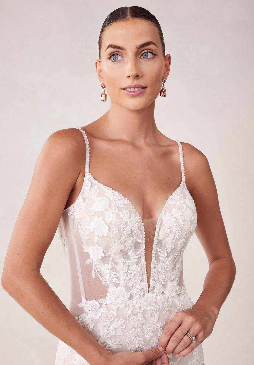 A woman with slicked-back hair wearing a white lace bridal gown with floral embroidery and thin straps, posing against a plain background.
