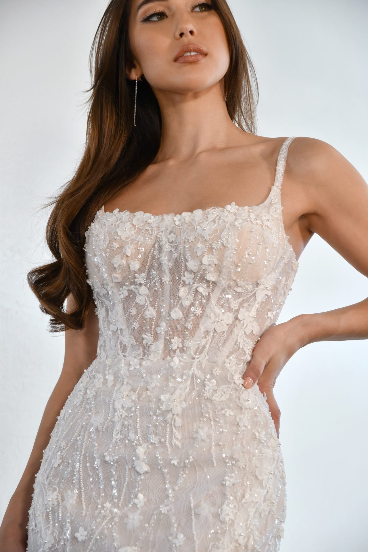 A woman wearing a white, intricately embroidered wedding dress with floral details, revealing her shoulder and part of her arm, with long, wavy brown hair and earrings, against a plain background.
