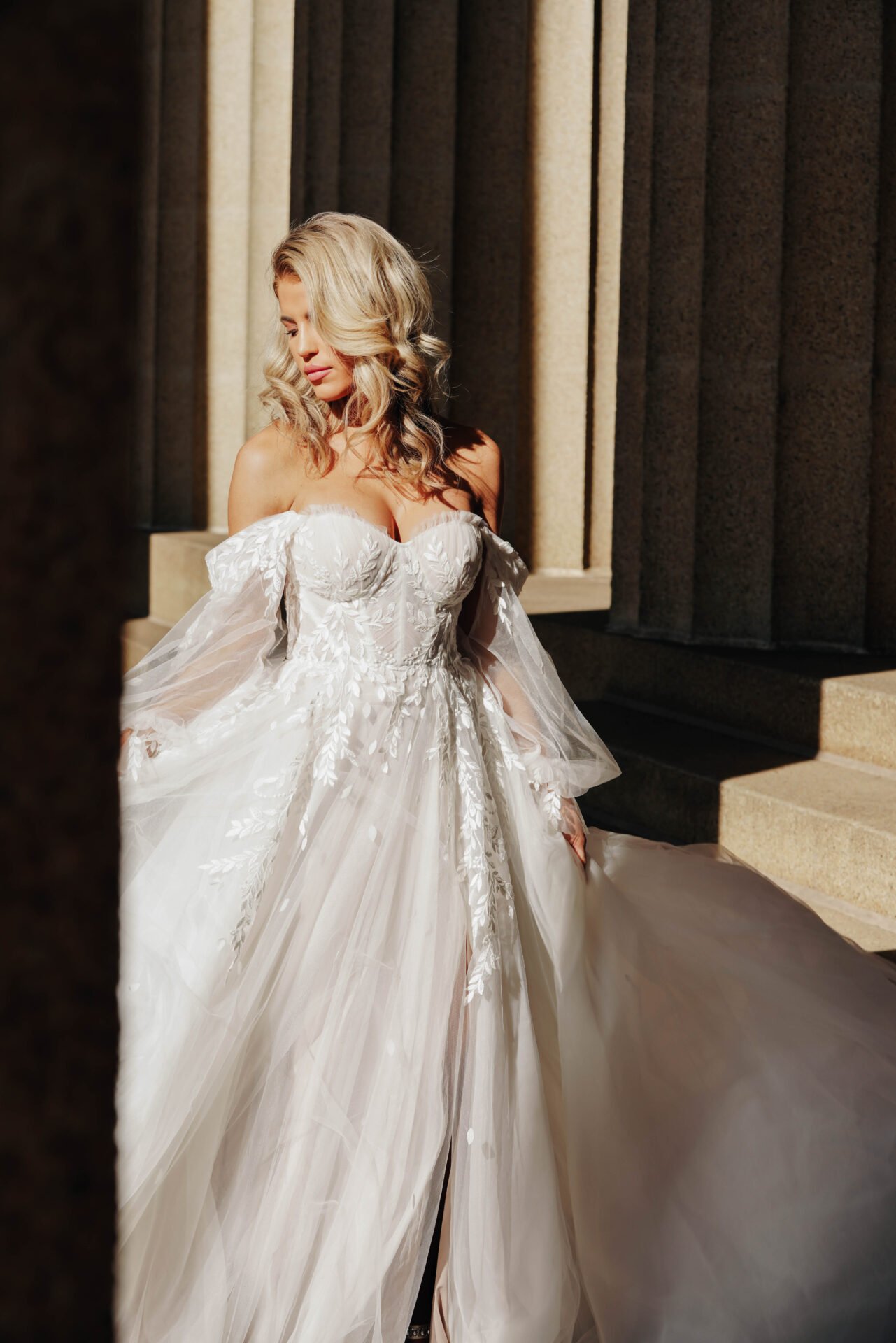 A woman in a wedding dress with off-the-shoulder sleeves and floral embroidery, standing outdoors near steps and stone columns.