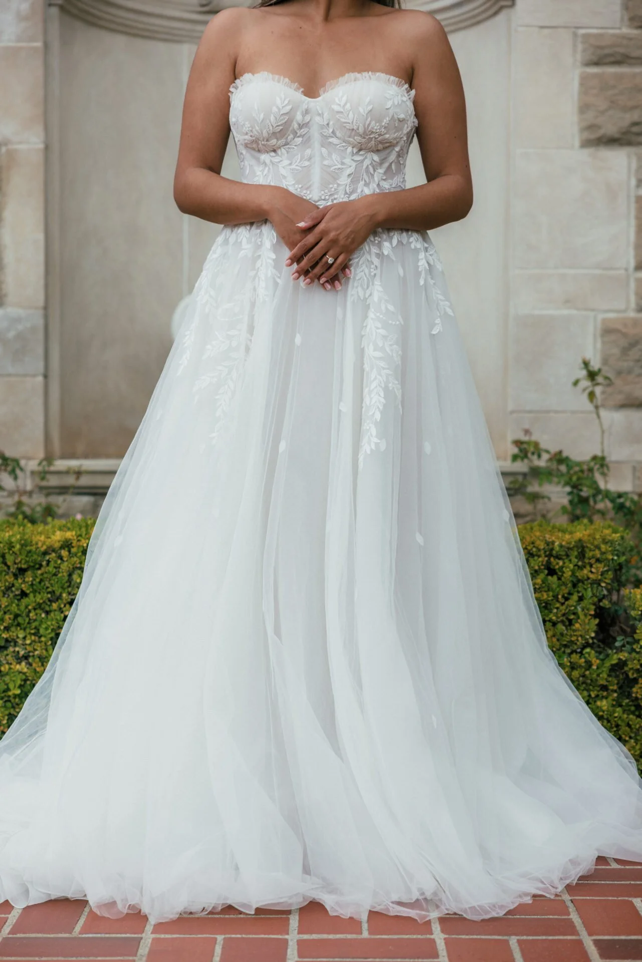 A woman wearing a strapless white wedding gown with floral embroidery, standing outdoors on a brick surface with a stone wall background.