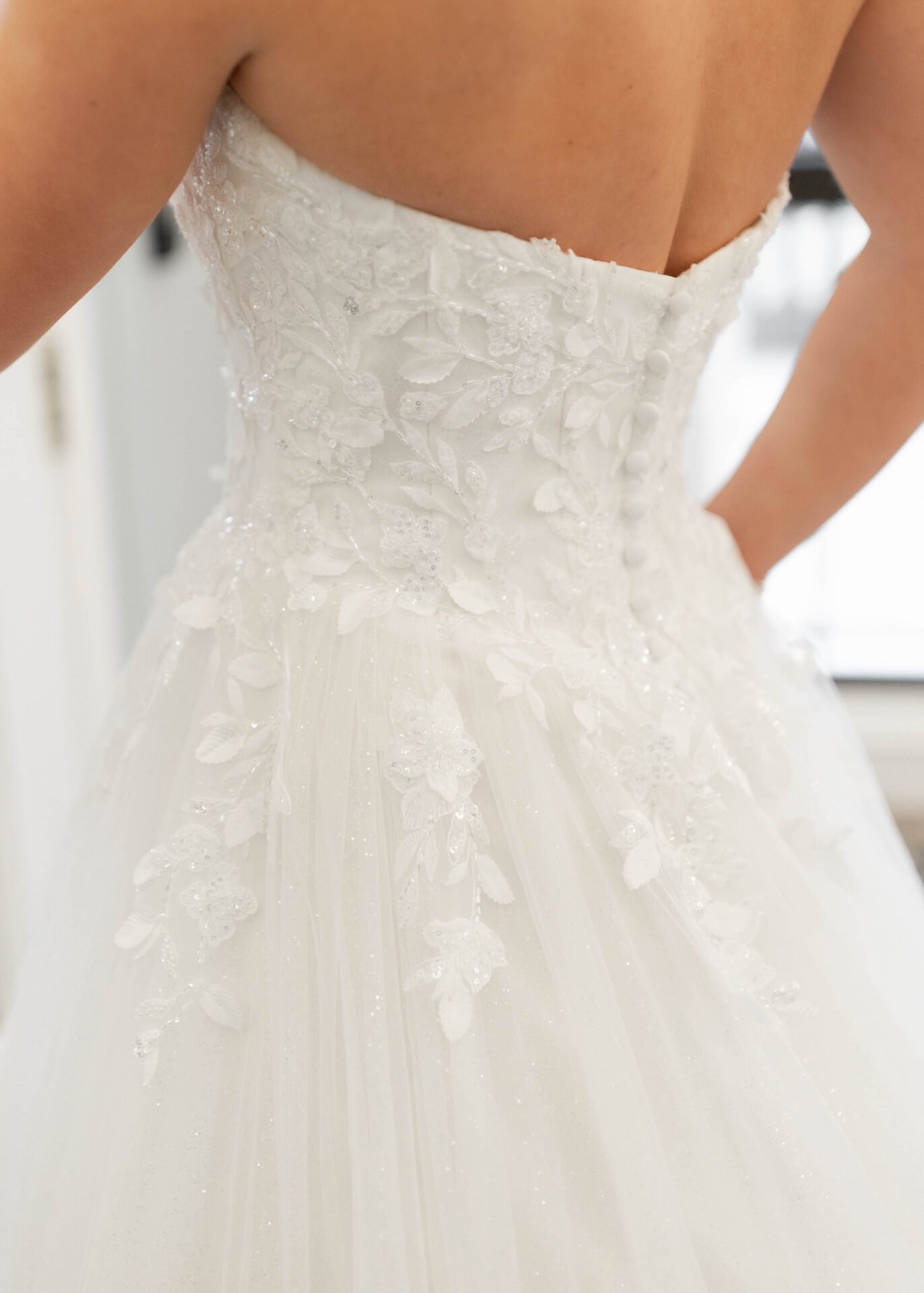 Close-up of a wedding dress with lace and floral embroidery, seen from the back with buttons down the center, worn by a person.