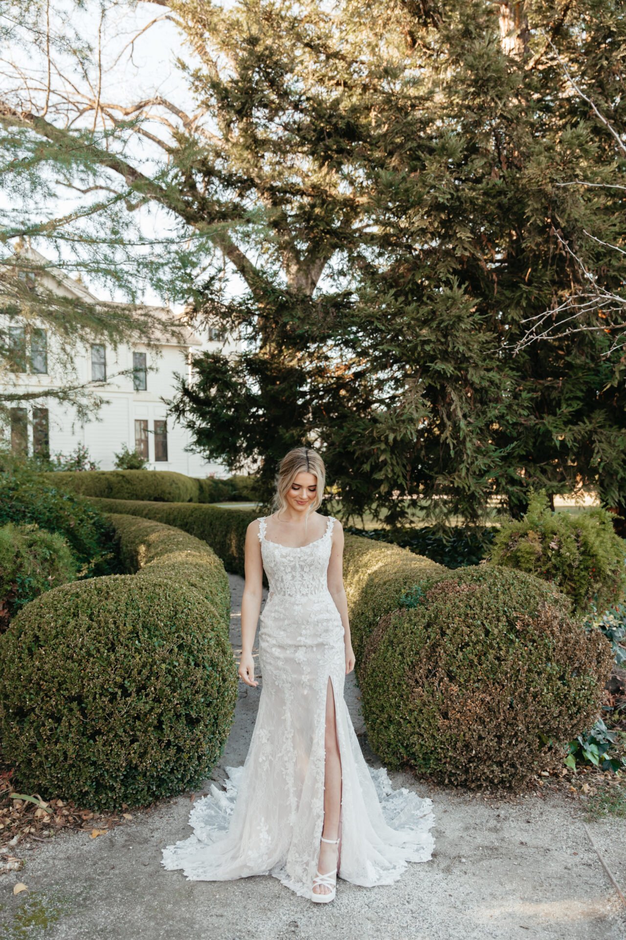 Bride in a lace wedding gown with a slit, standing on a garden path lined with trimmed bushes and tall trees, smiling softly.