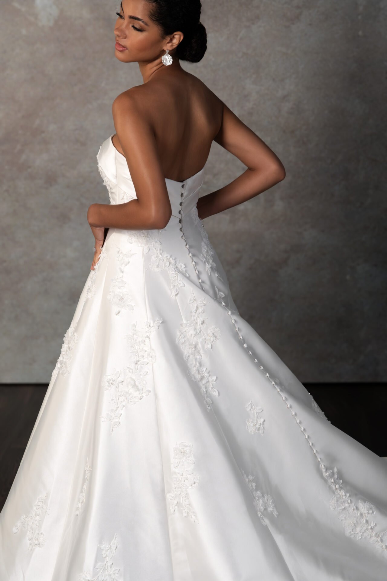 A woman wearing a white strapless wedding gown with lace embroidery, posing with her back partly turned, against a simple textured background.