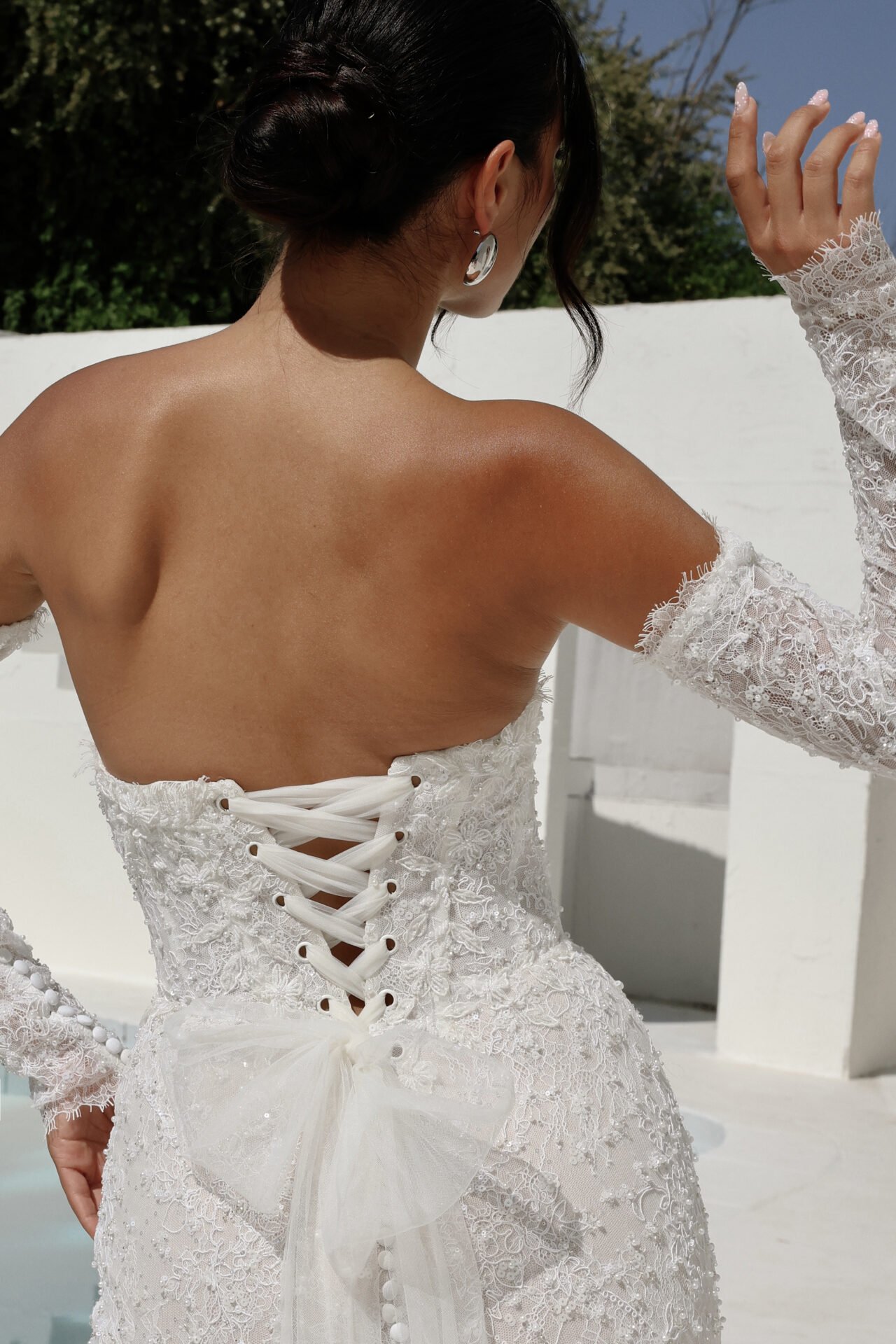 Back view of a woman wearing a white lace wedding dress with a corset back and bow, outdoors under a clear blue sky.