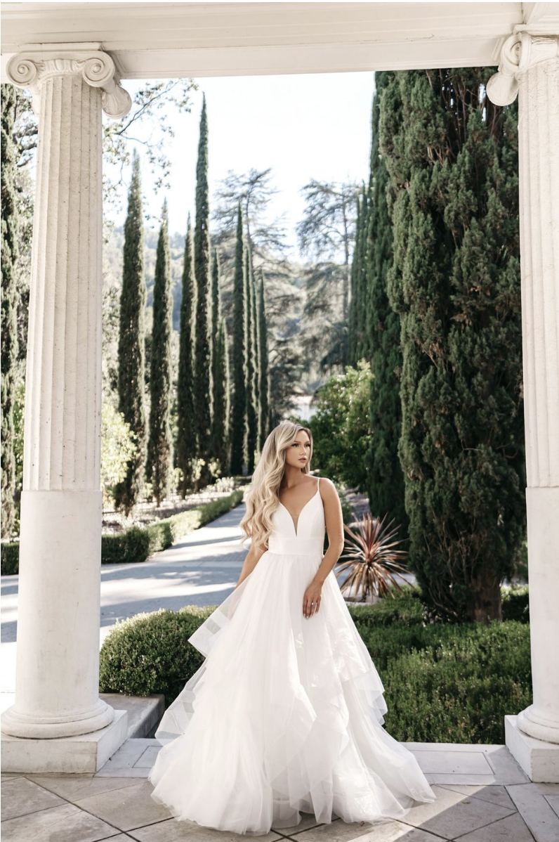 A woman in a white wedding dress standing between white classical columns, with a backdrop of tall cypress trees and greenery.