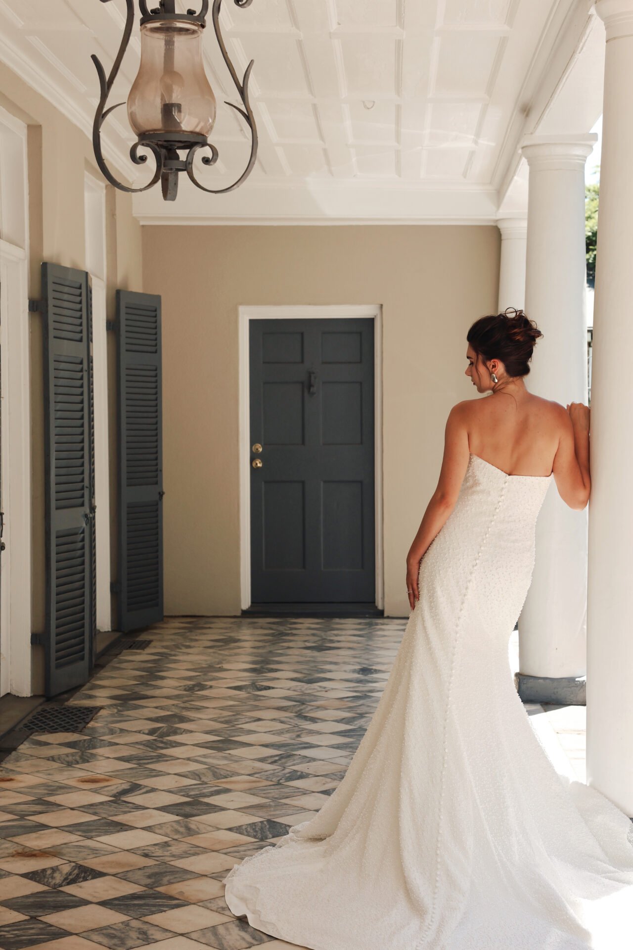 A woman in a strapless wedding dress standing on a patterned tile floor, leaning against a white column in an entryway, with a dark door and window shutters in the background.