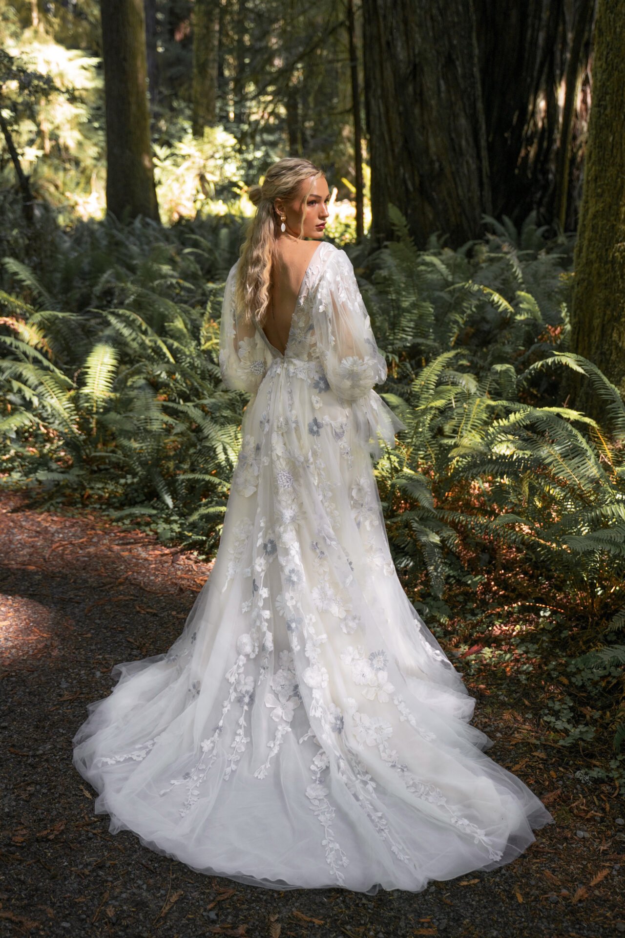 A woman wearing a white, embroidered wedding dress with long sleeves, standing in a lush forest, looking to her right.