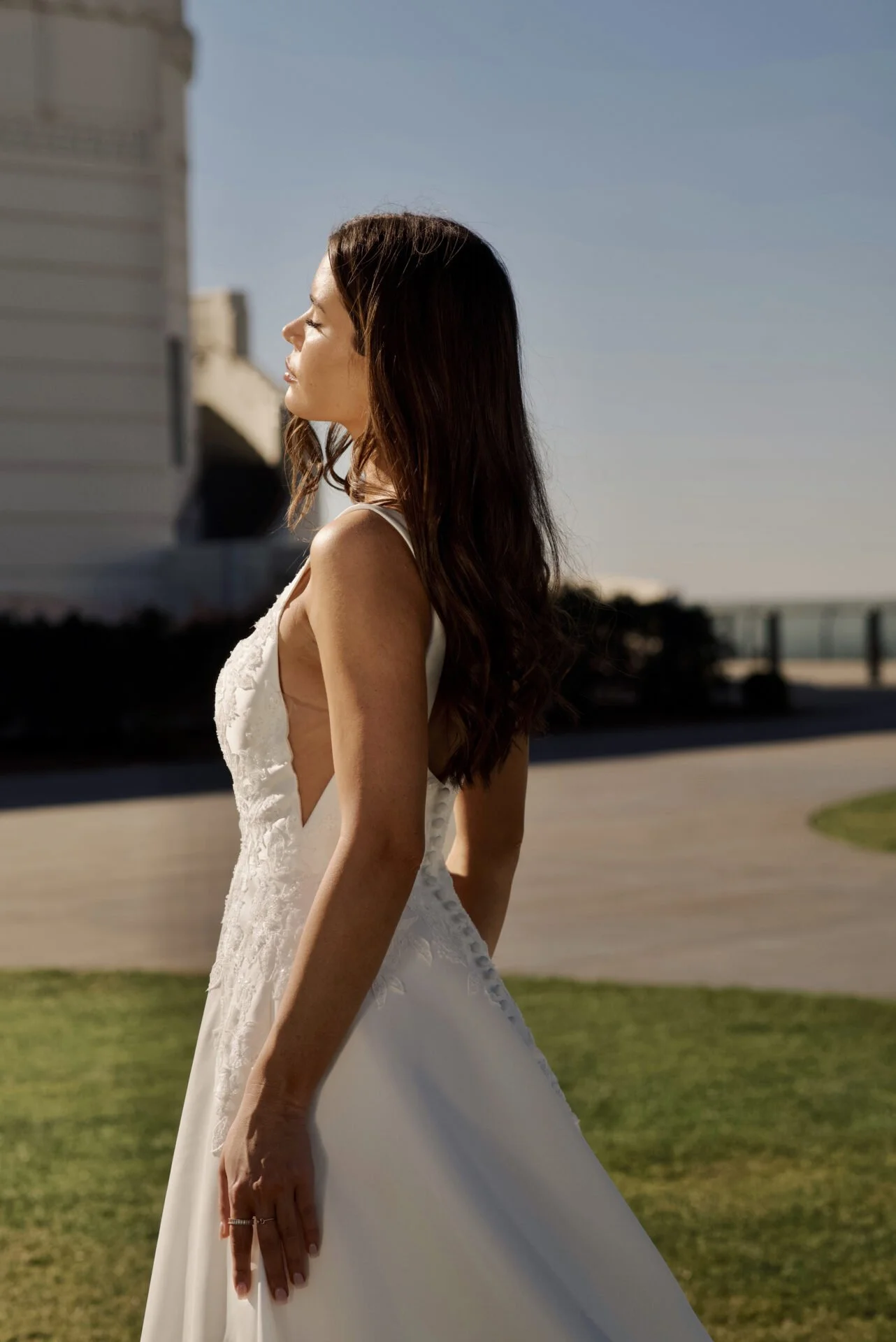 A woman in a wedding dress with long, wavy brown hair stands outdoors with her eyes closed and face turned slightly to the side, basking in sunlight.