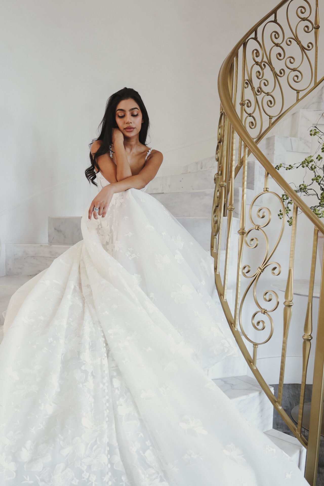 A woman in a white wedding dress standing on a marble staircase next to a gold ornate railing.