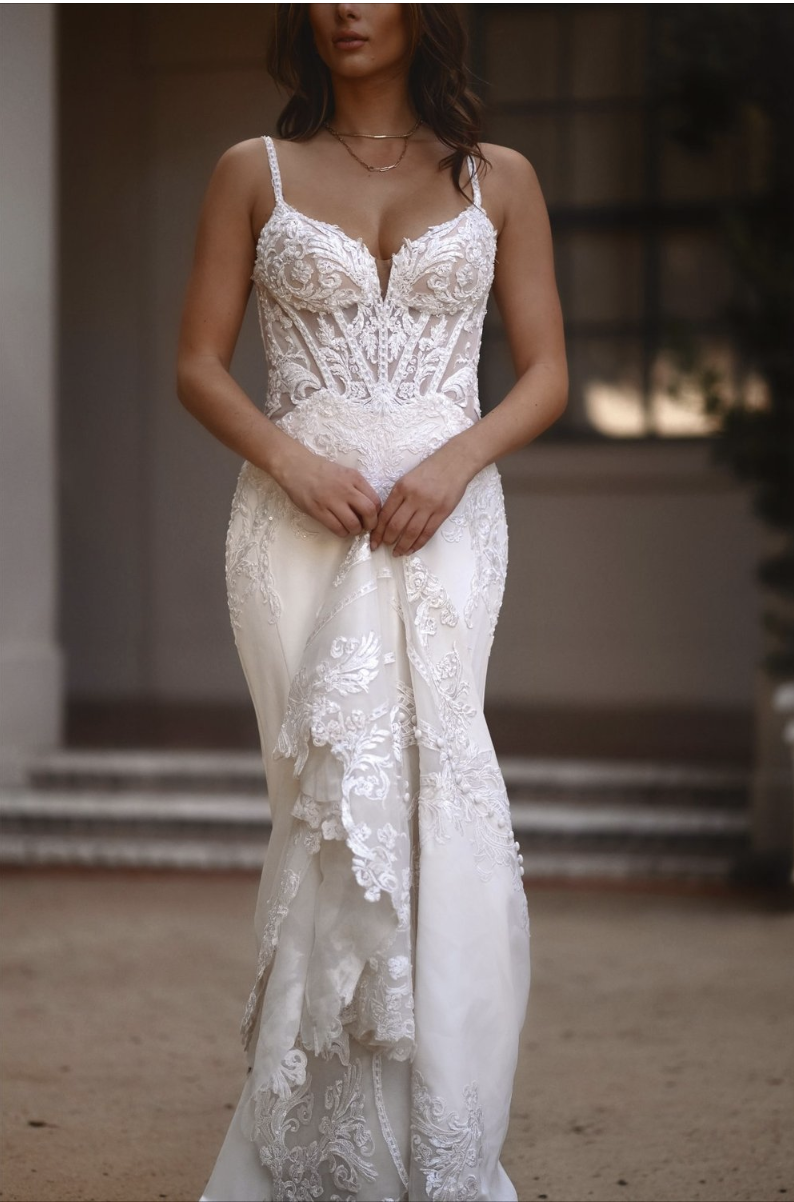 Woman wearing a white lace wedding dress with spaghetti straps, standing outdoors with a blurred background.