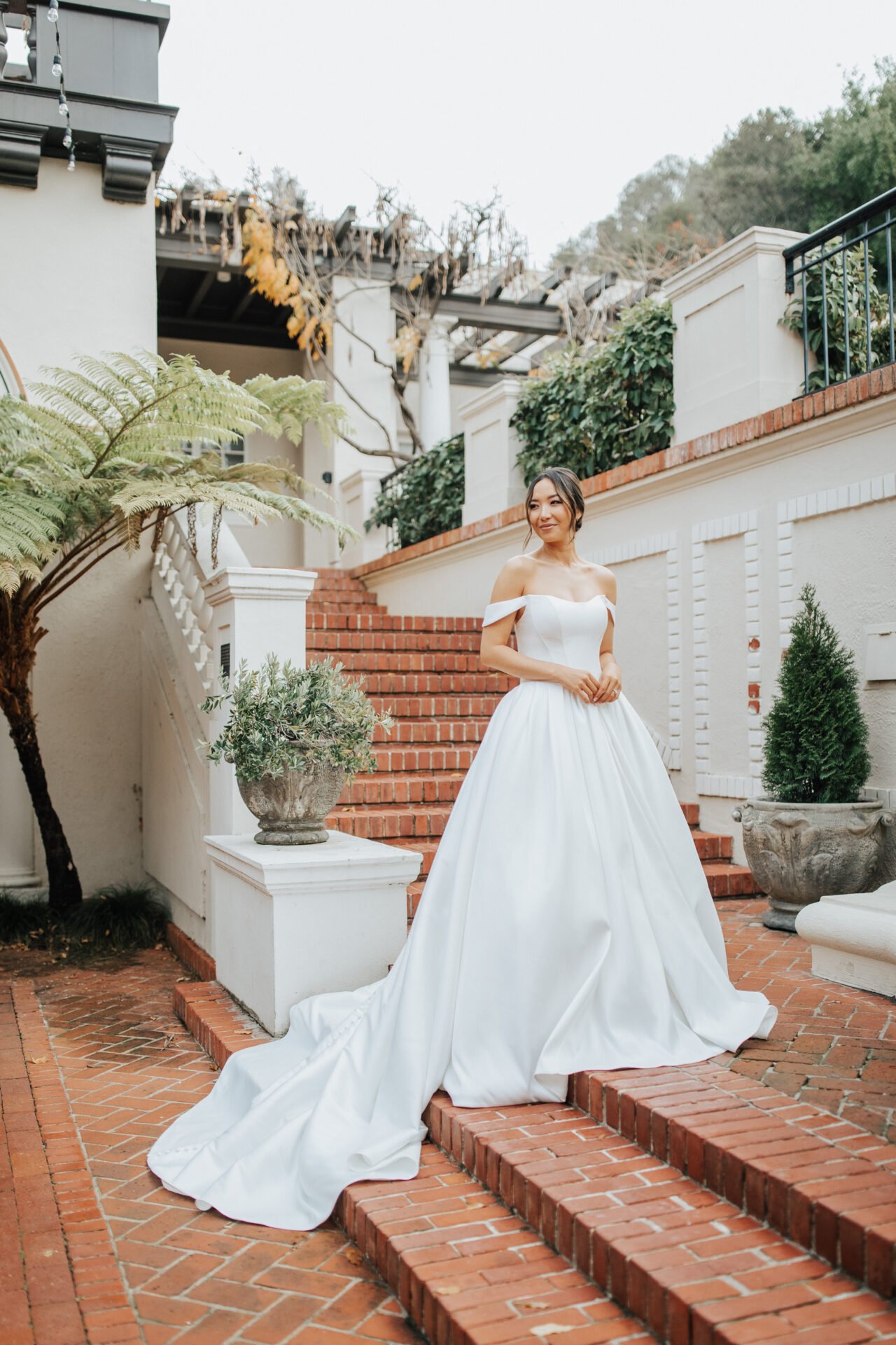 A woman in a white off-shoulder wedding gown standing on brick stairs outside a white building with greenery and potted plants.