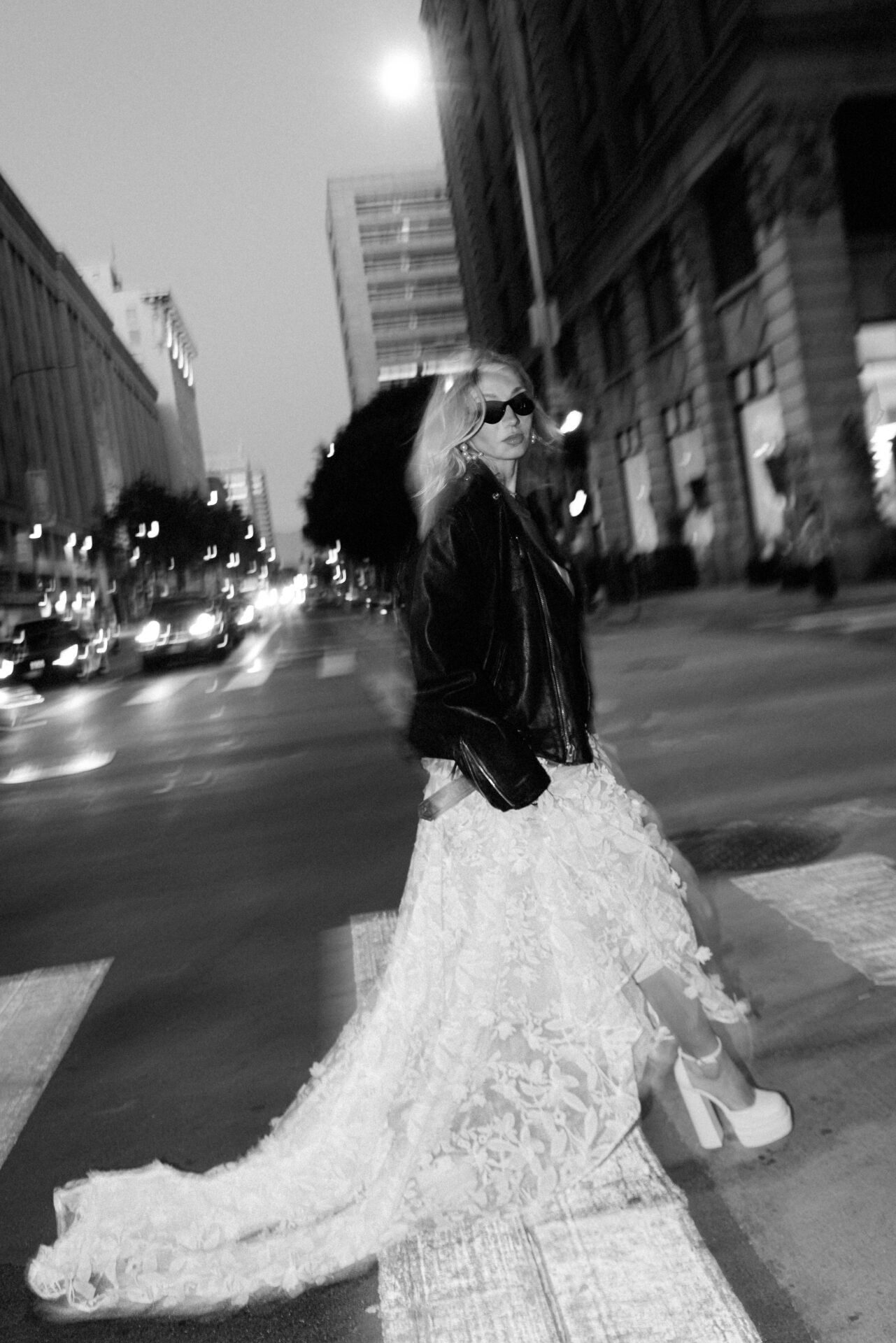 A woman with blonde hair wearing sunglasses, a black leather jacket, and a long lace dress, walking on a city street at night.
