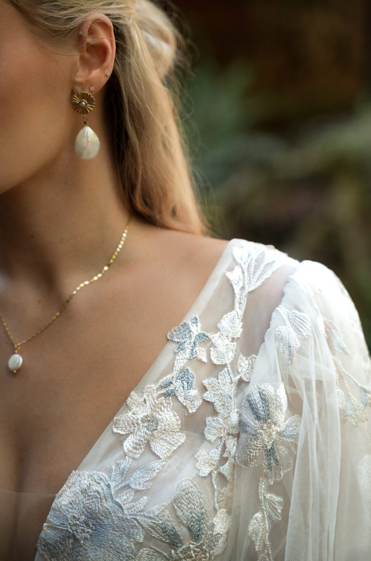 Close-up of a woman wearing a white lace dress with floral embroidery, gold necklace with a pearl pendant, and a gold flower-shaped earring with a teardrop-shaped pearl, set against a blurred outdoor background.