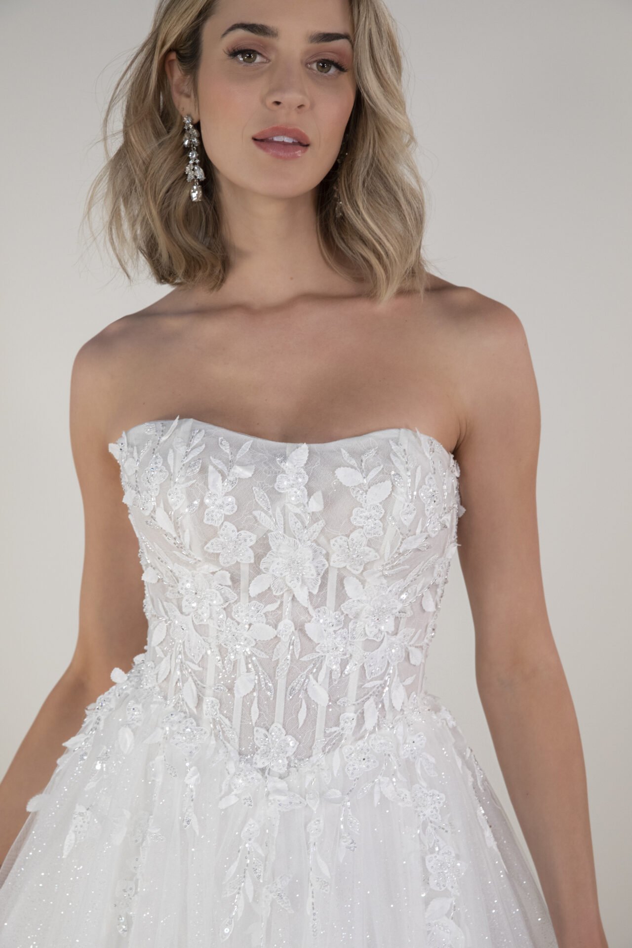 A woman wearing a strapless white wedding dress with floral embroidery and lace details, and dangling earrings.