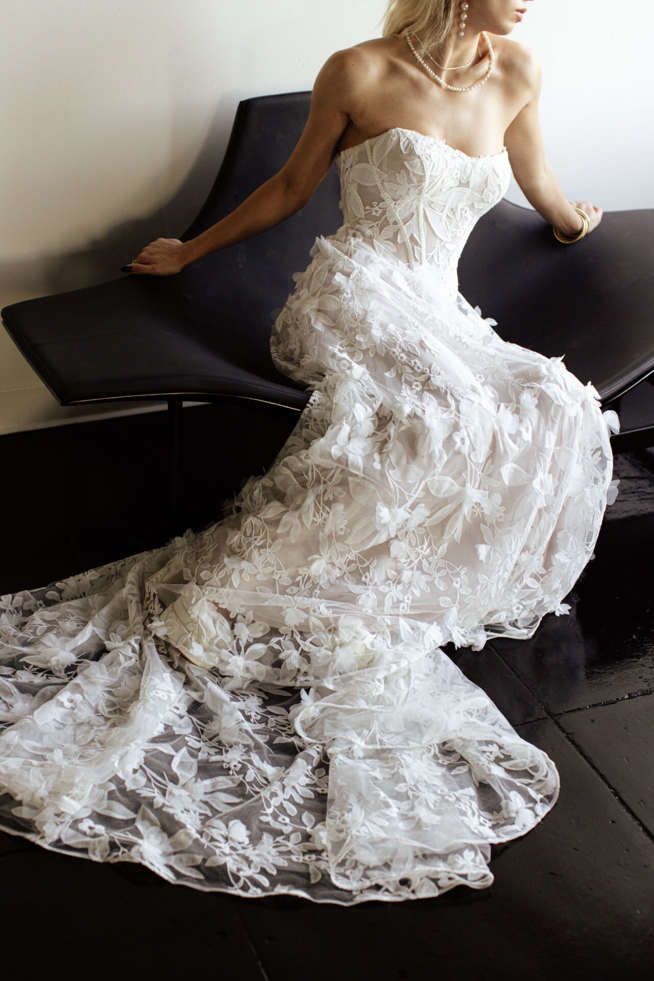 Woman wearing a strapless white lace wedding dress and pearl jewelry sitting on a black bench.