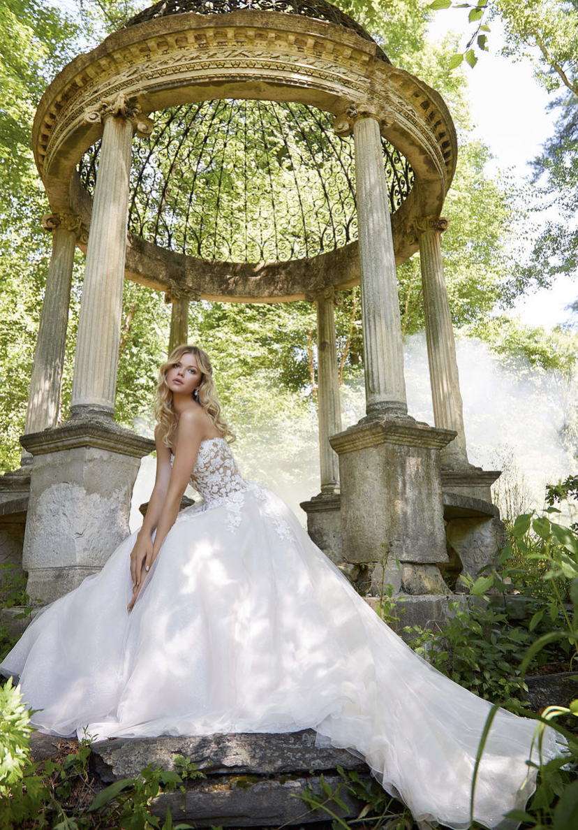 A woman in a white wedding dress with lace details sitting on stone steps in front of an ornate stone and metal gazebo in a lush green garden.