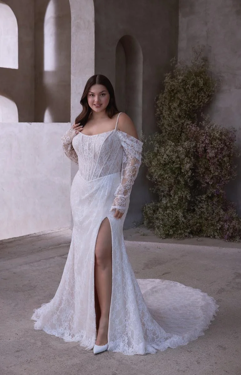 A woman in a white lace wedding dress with off-shoulder sleeves and a high slit, standing on a neutral floor against a background of textured gray walls and greenery.