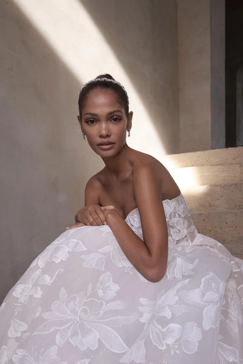 A young woman with dark skin and short hair styled in a bun, wearing a strapless white dress with floral embroidery, sitting on stairs against a plain wall, with sunlight casting a shadow across her face and upper body.