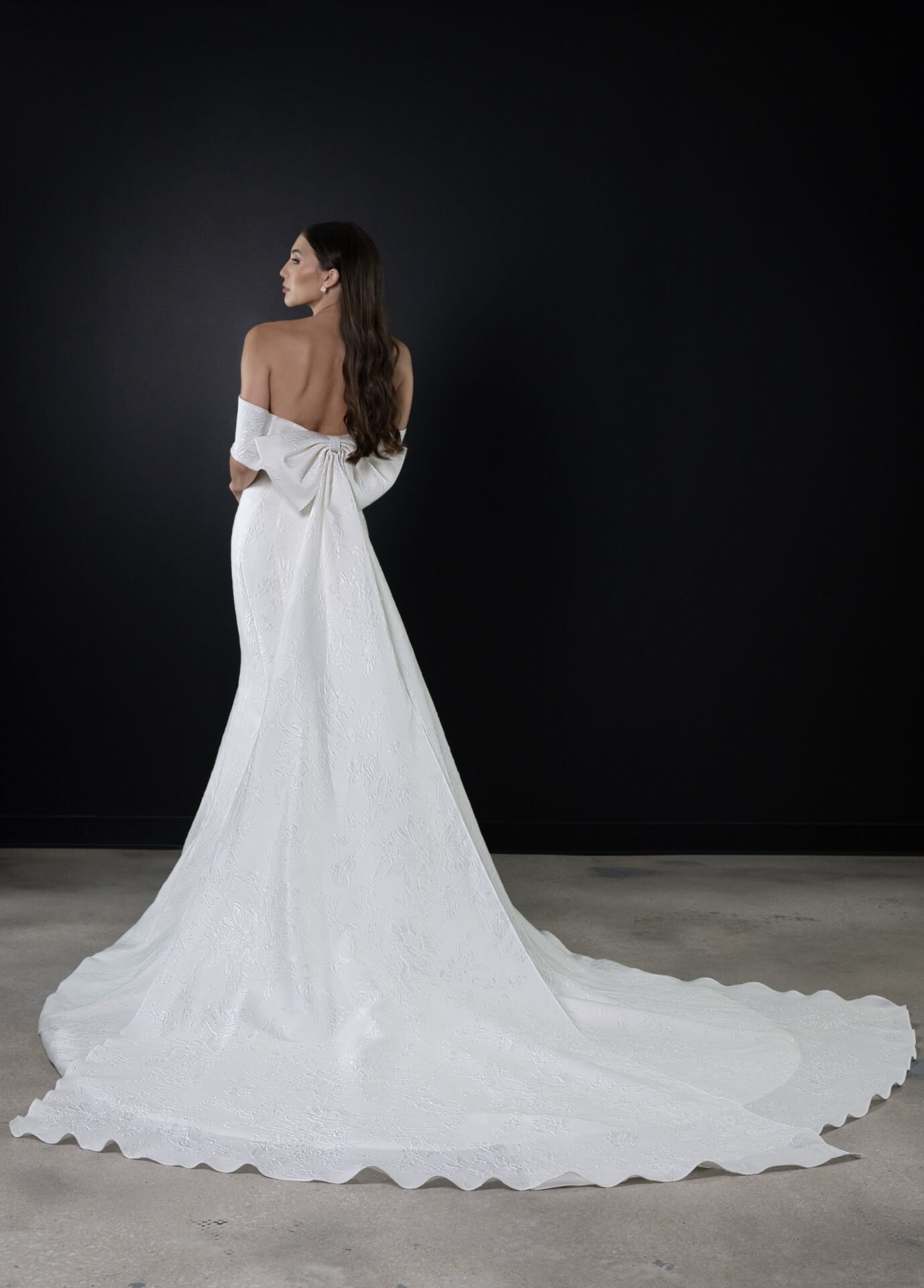 Woman wearing an elegant white wedding dress with a large bow in the back, standing against a dark wall.