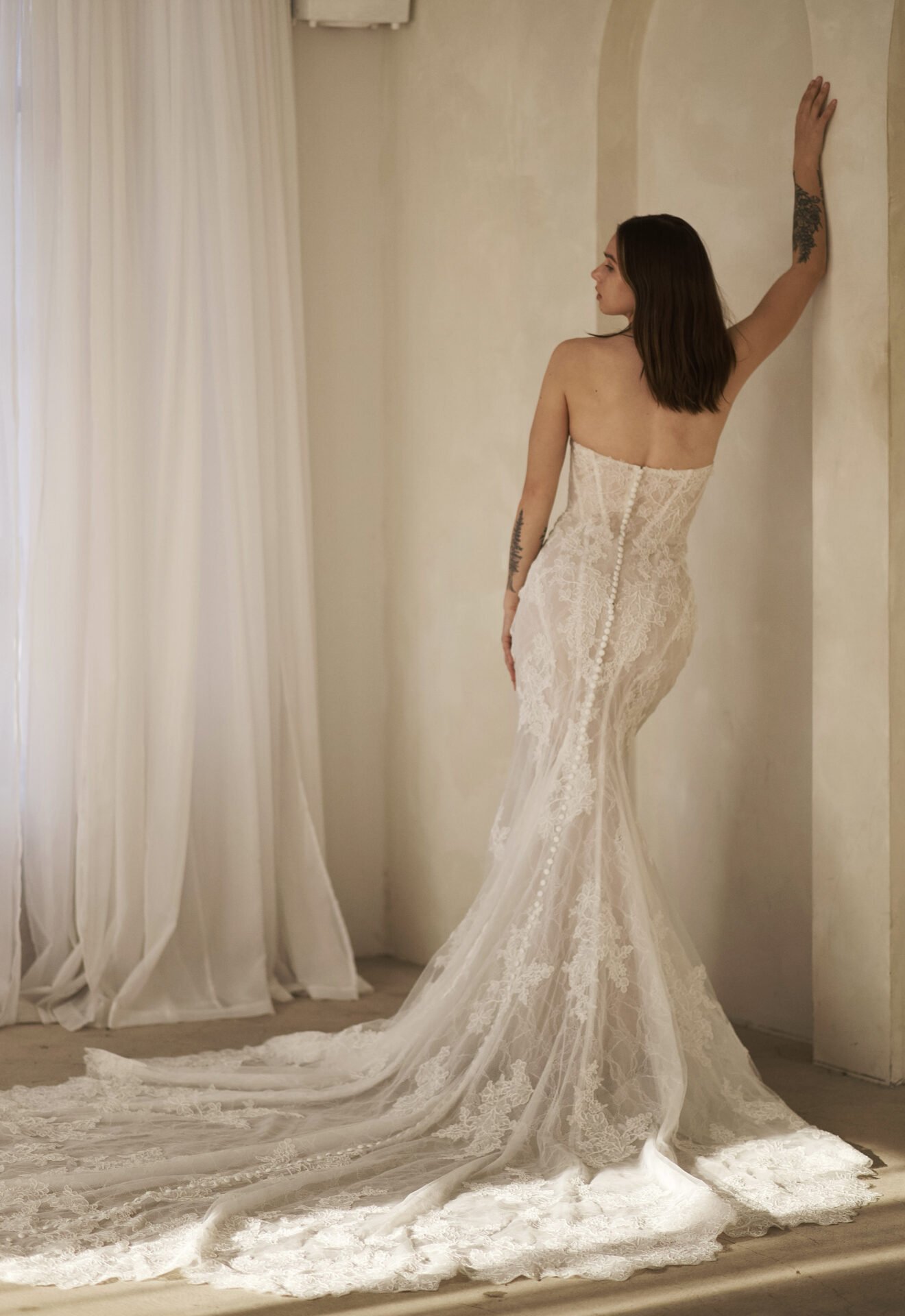 Woman in a white lace wedding dress standing with her back to the camera, one hand on the wall, in a softly lit room with cream-colored curtains.