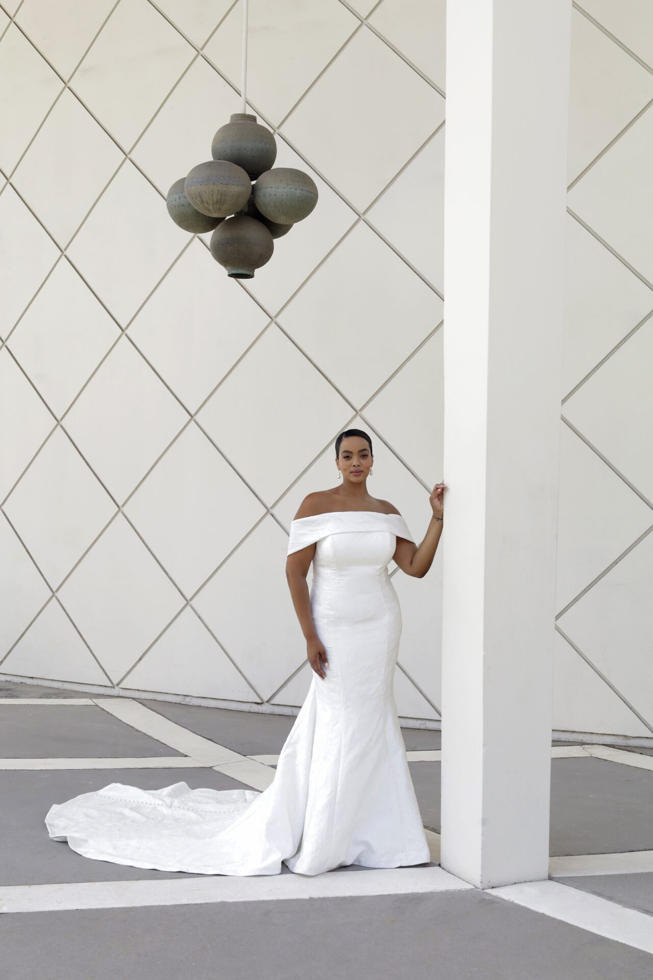 A woman in a white off-the-shoulder wedding gown standing beside a white pillar in front of a modern white wall with a geometric pattern, with a black hanging light fixture above.