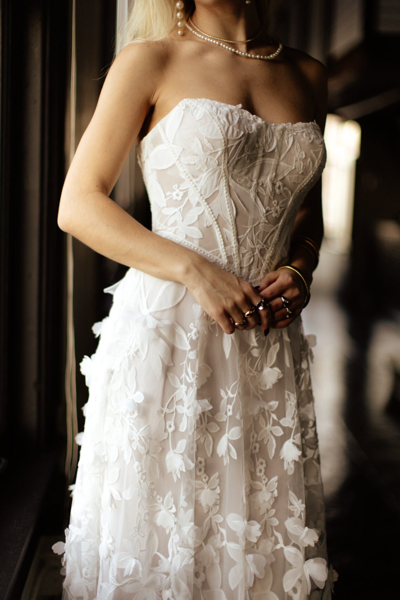 Close-up of a woman in a strapless white embroidered wedding dress with floral patterns, accessorized with pearl necklaces and rings.