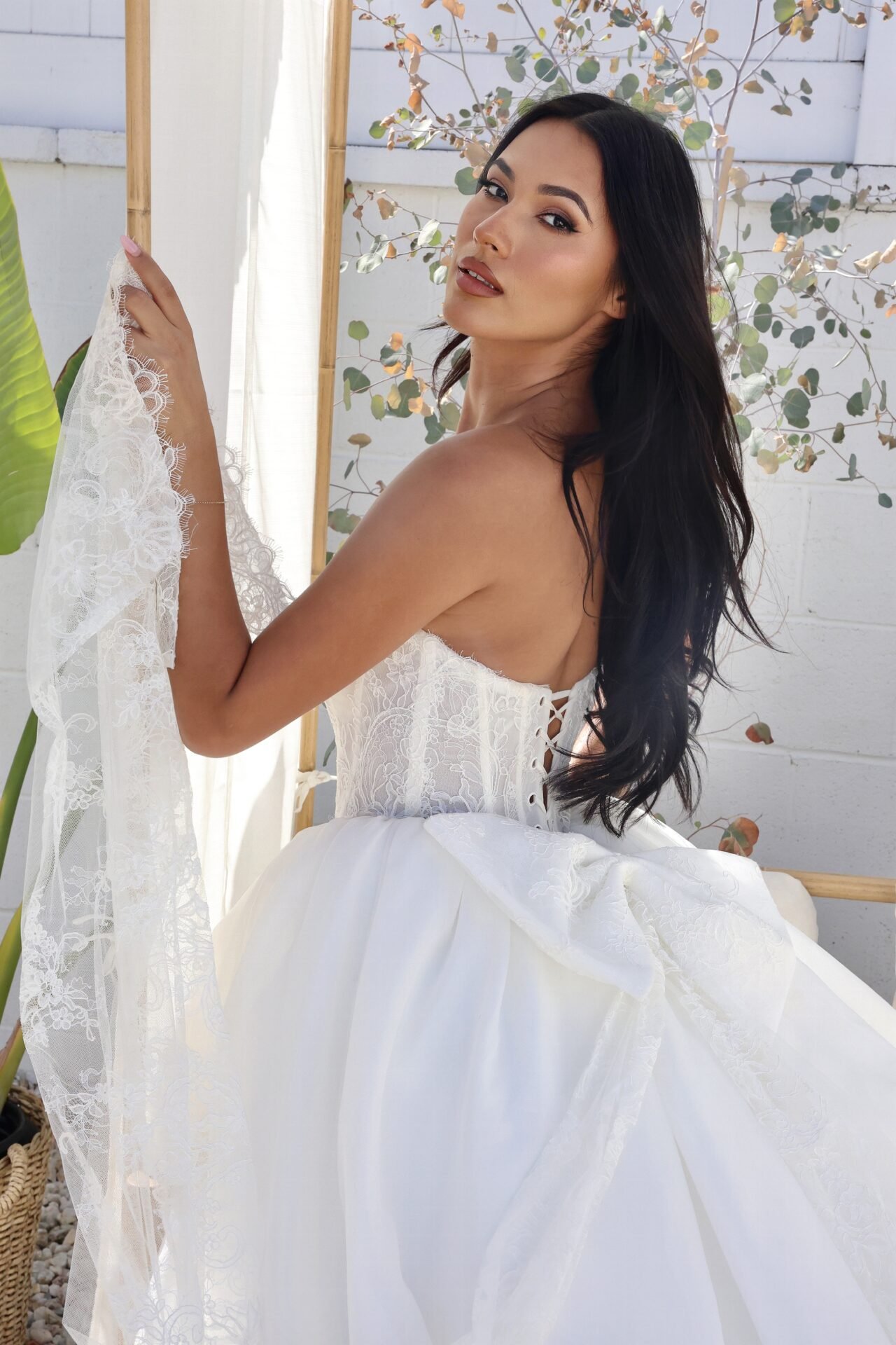 A woman with long black hair wearing a white dress with lace and corset details, sitting outdoors amid plants and a white wall, looking over her shoulder.