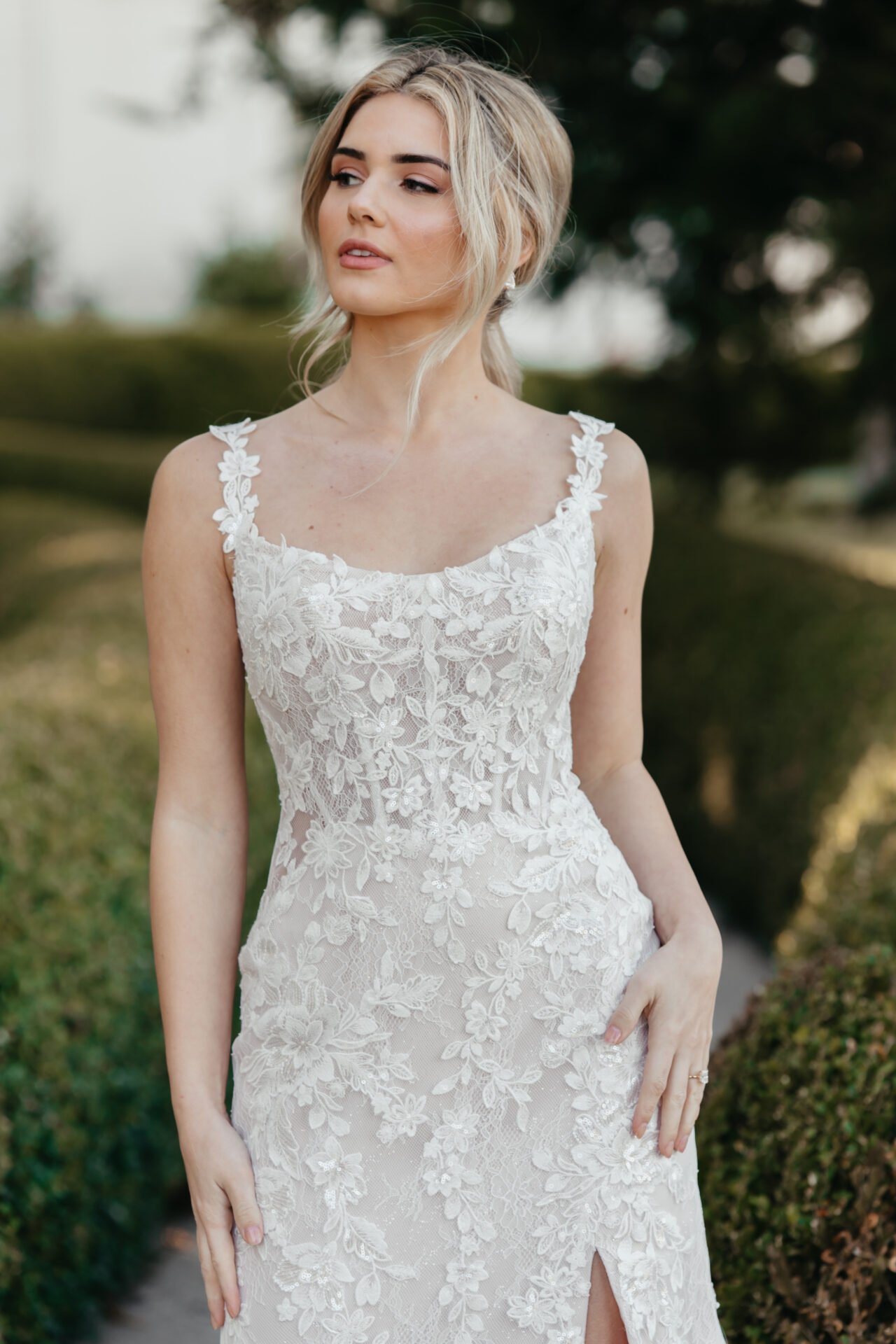 A young woman in a white lace wedding dress standing outdoors with trees and greenery in the background.