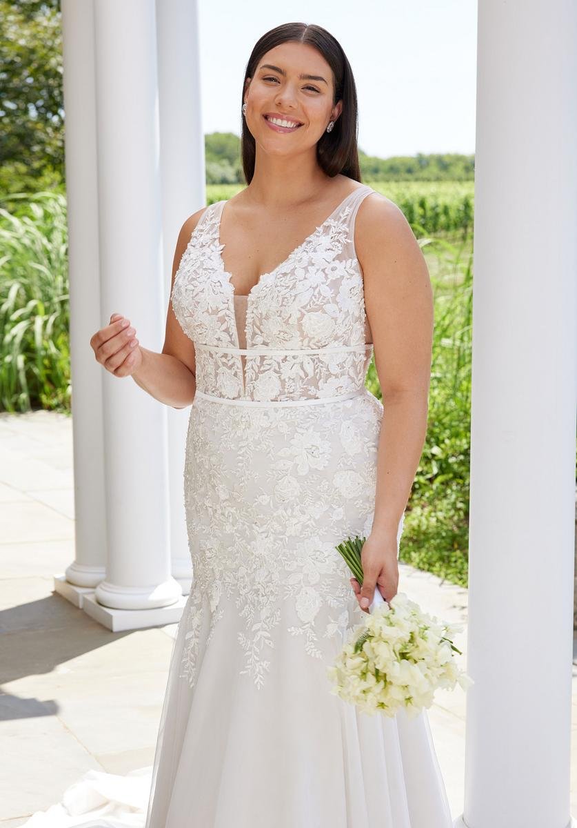 A smiling woman in a white lace wedding dress holding a bouquet of white flowers, standing outdoors near white columns with greenery in the background.