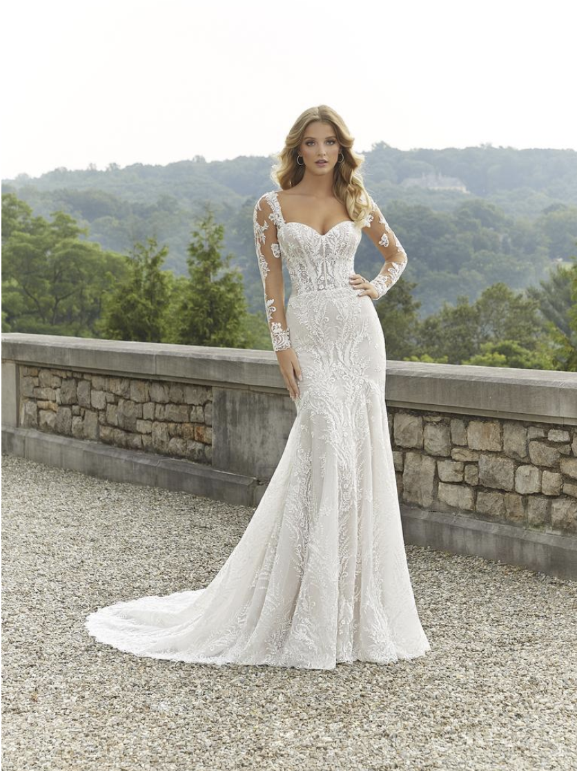 A woman in a white, long-sleeved, lace wedding gown with a fitted bodice and a train, standing outdoors near a stone wall with a scenic, green, hilly landscape in the background.
