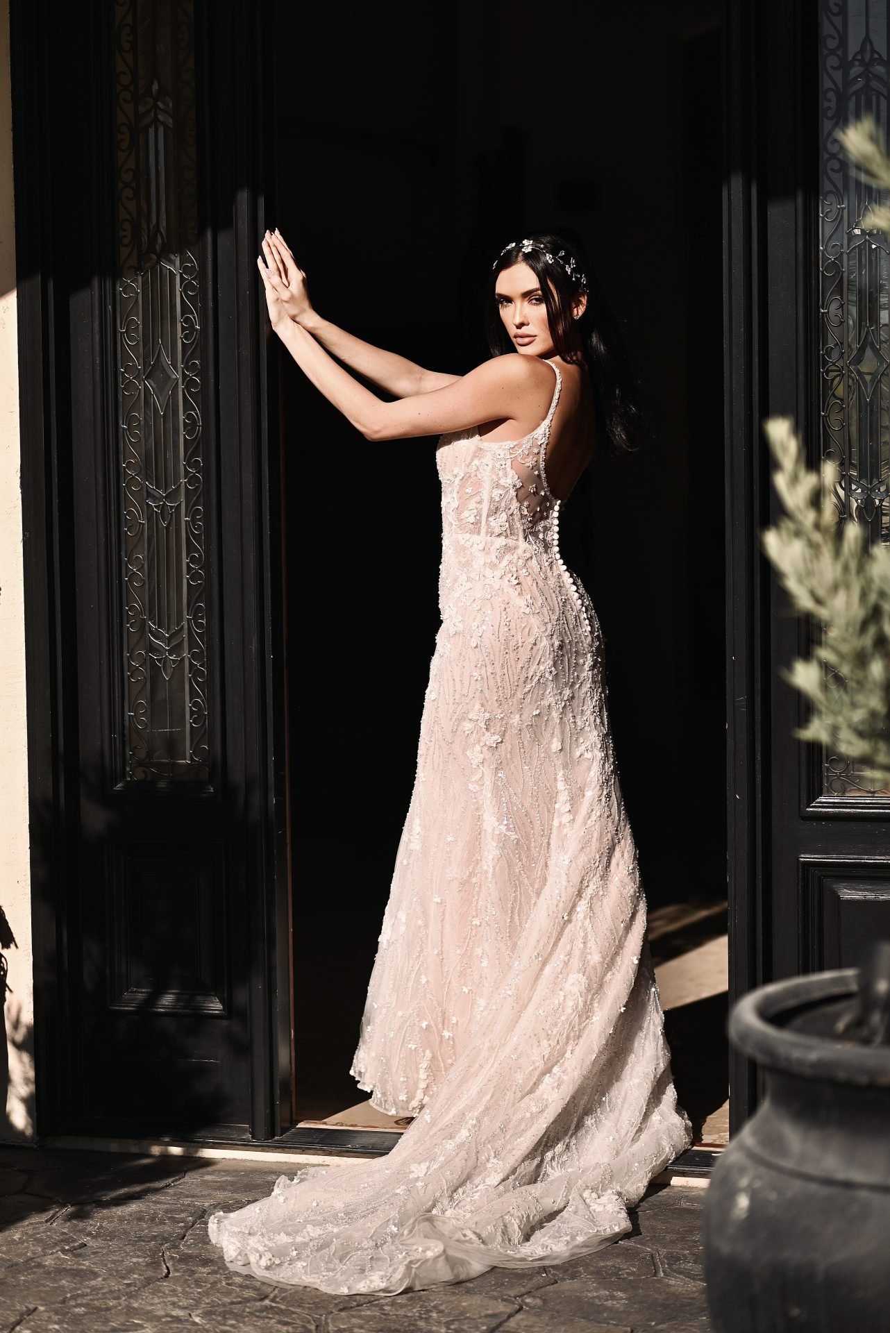 A woman in a wedding dress standing in a doorway, posing with her arms extended, with sunlight highlighting her dress and dark background.