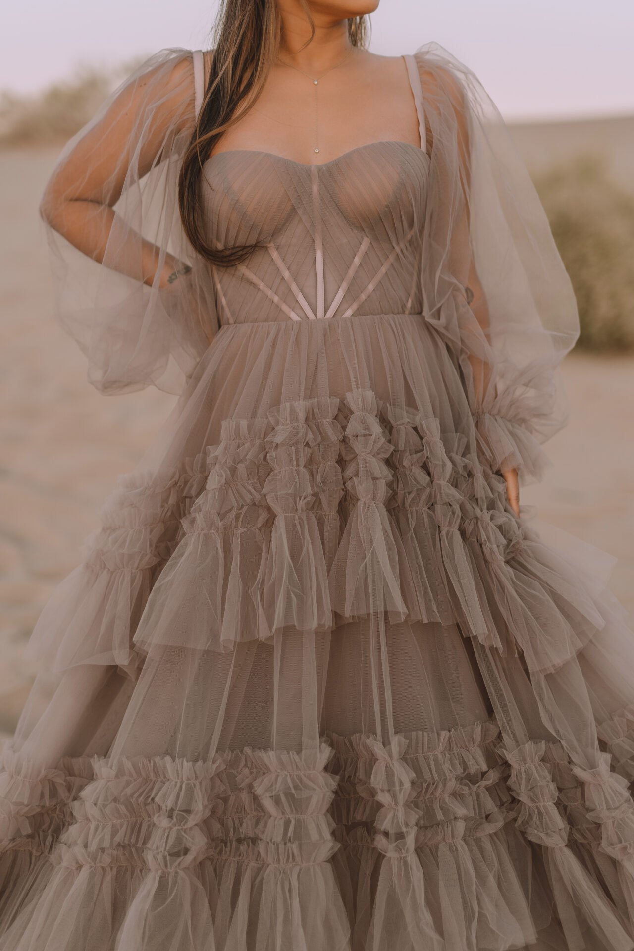 A woman wearing an elaborate nude-colored tulle dress with ruffles and a fitted bodice, standing outdoors on a sandy landscape during sunset.