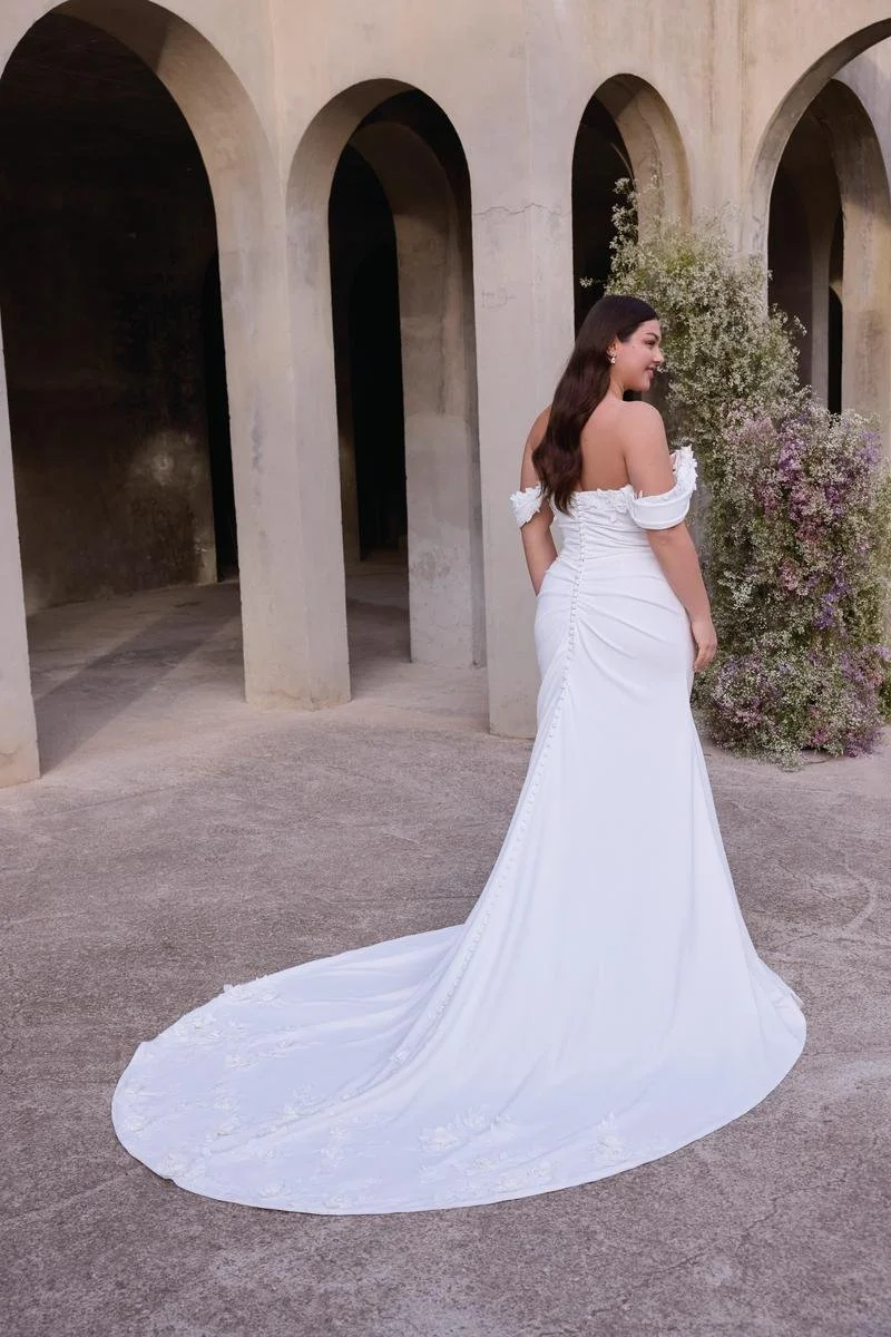 A woman in a white wedding dress standing outdoors near a wall with arched openings and a flowering plant in the background.