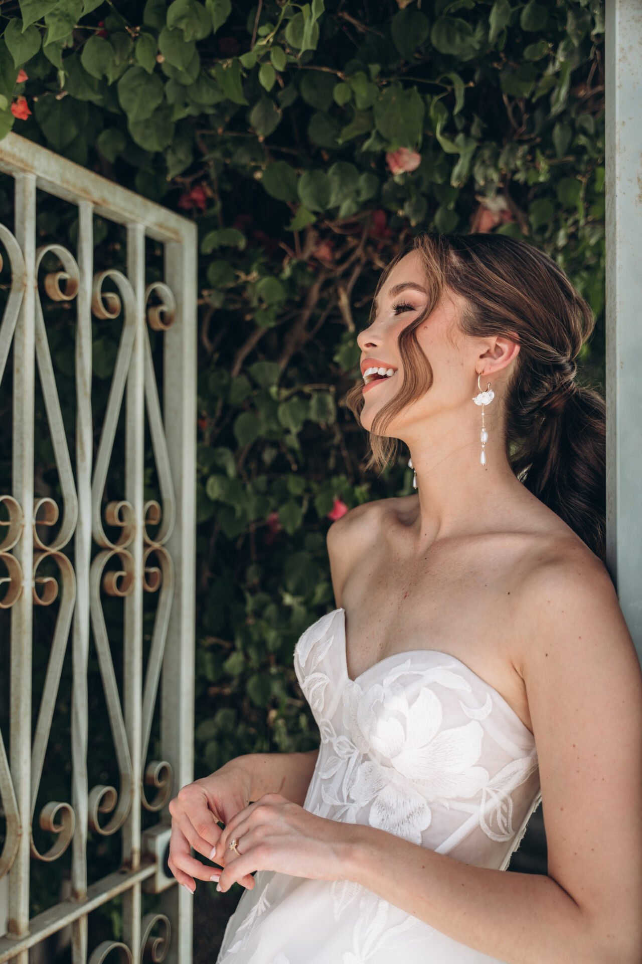 A woman in a white wedding dress smiling and looking to her right, standing outdoors near a rusty metal gate and green leafy bushes.
