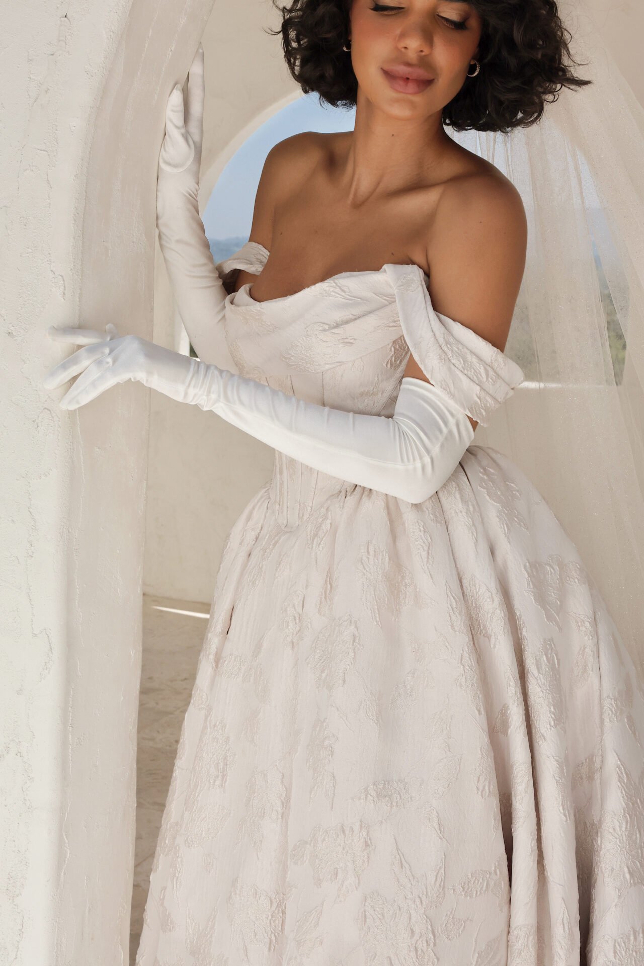 A woman in an elegant off-shoulder wedding dress with long gloves posing near a white wall.