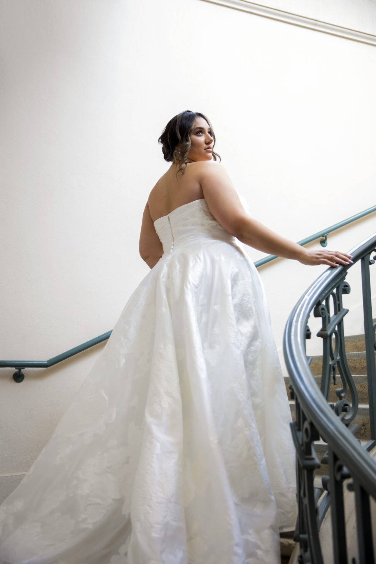 A woman in a white strapless wedding gown standing on a staircase, holding the railing, looking to the side.