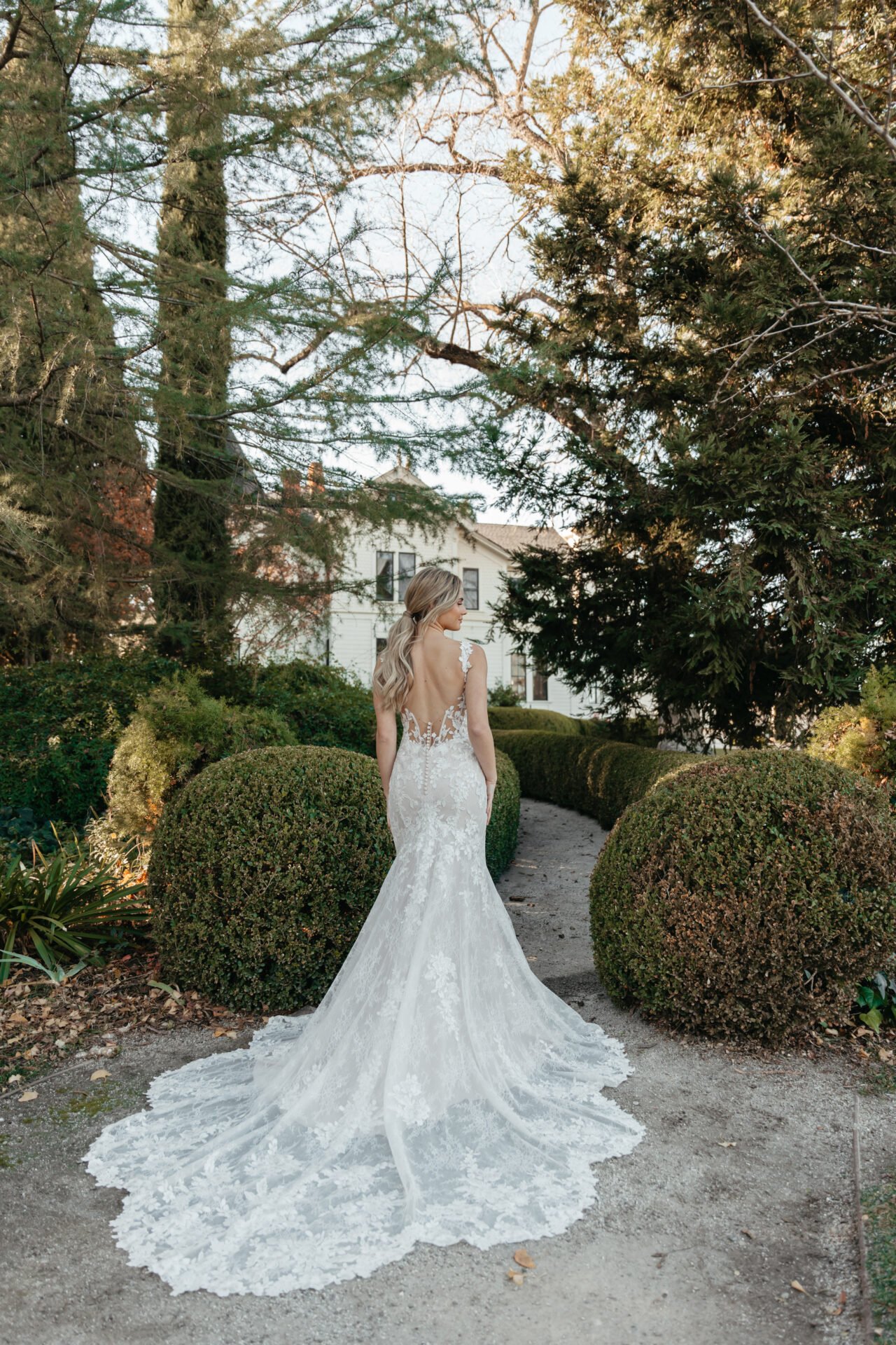 Bride in a white lace wedding gown with an open back and long train standing on a garden path surrounded by bushes and trees.