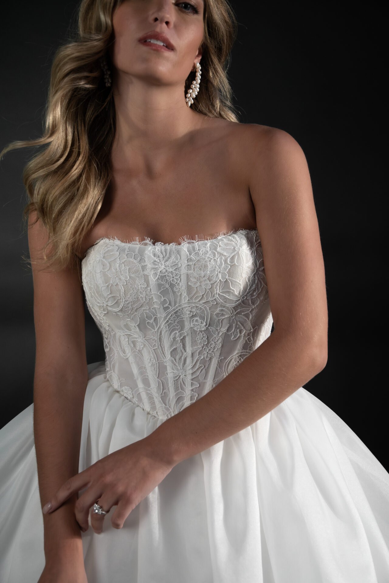 Woman wearing a strapless white wedding dress with intricate lace embroidery, large pearl earrings, and a ring on her left hand against a dark background.