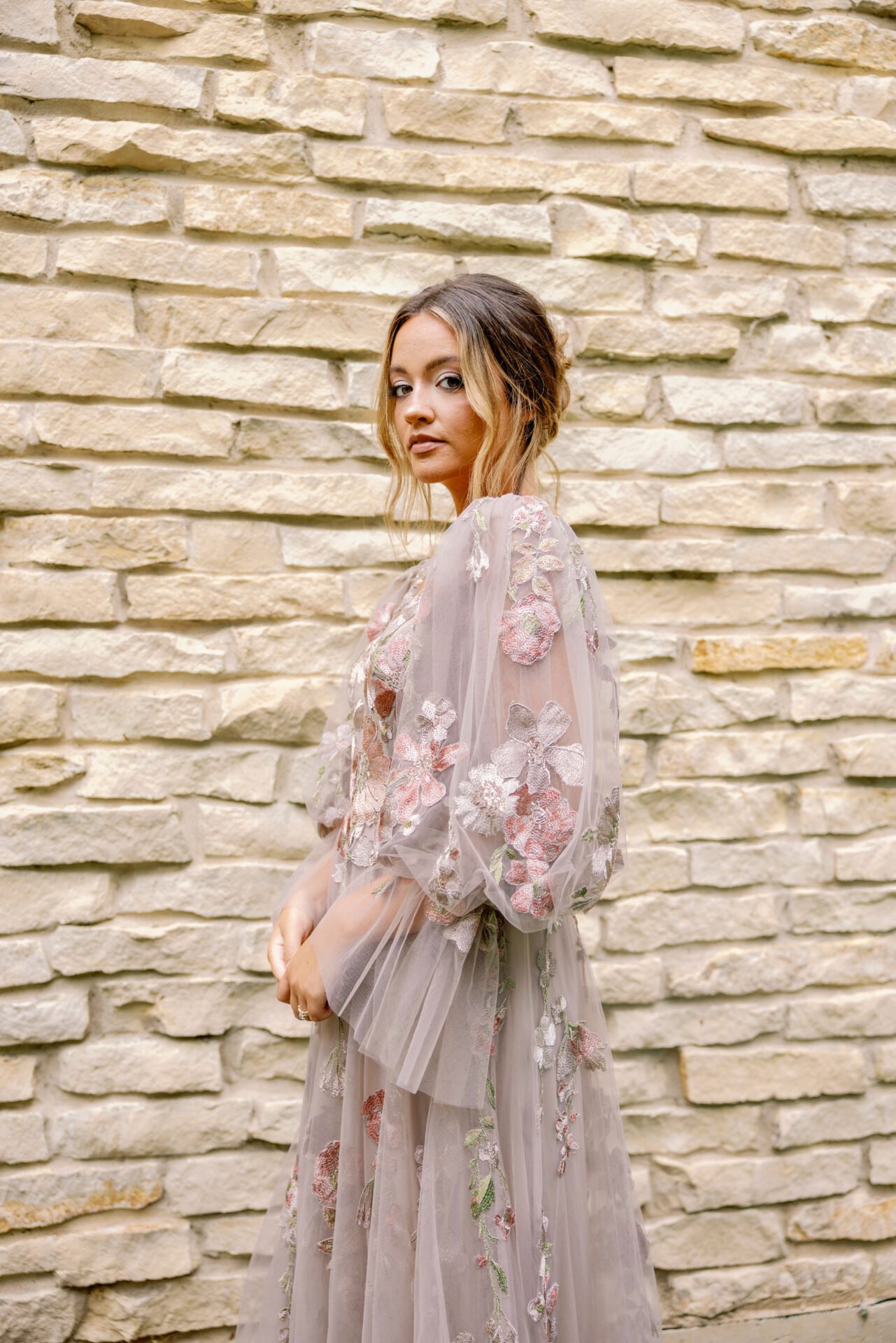 A woman wearing a sheer, embroidered floral gown standing in front of a cream-colored stone wall.