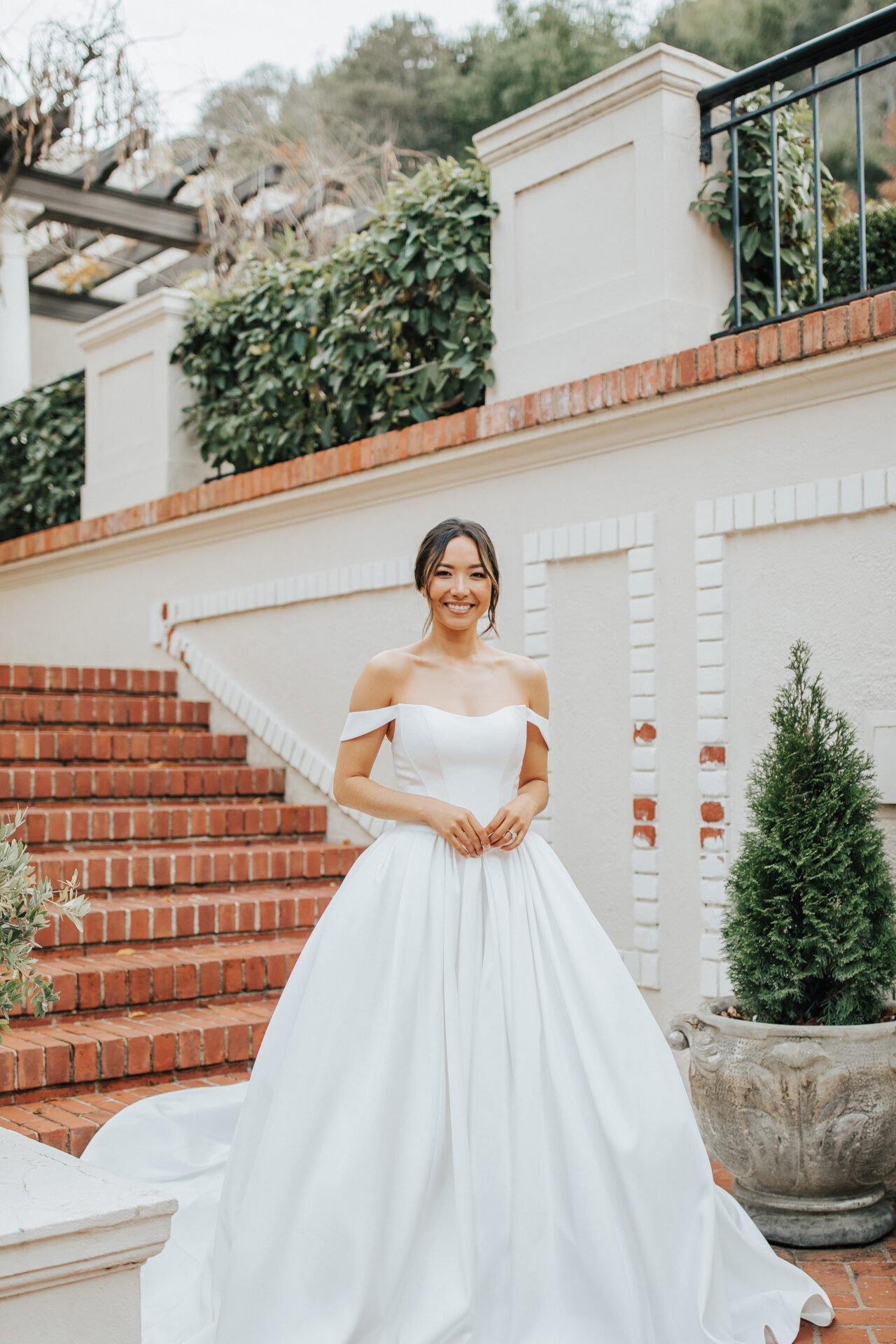 Woman in a white off-shoulder wedding dress standing on brick steps outside, smiling, with greenery and a white wall in the background.