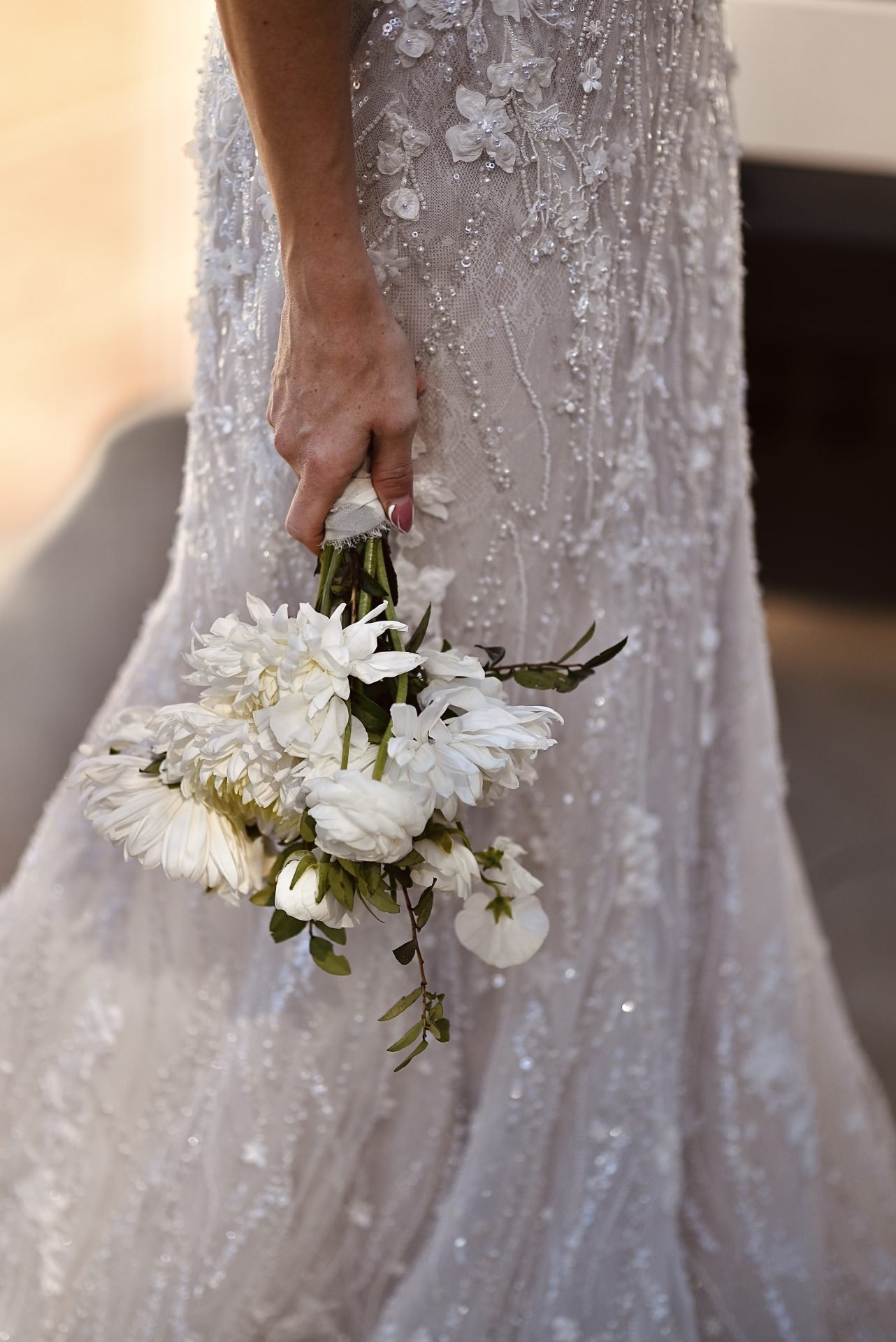 Close-up of a bride in a detailed, beaded wedding dress holding a small bouquet of white flowers