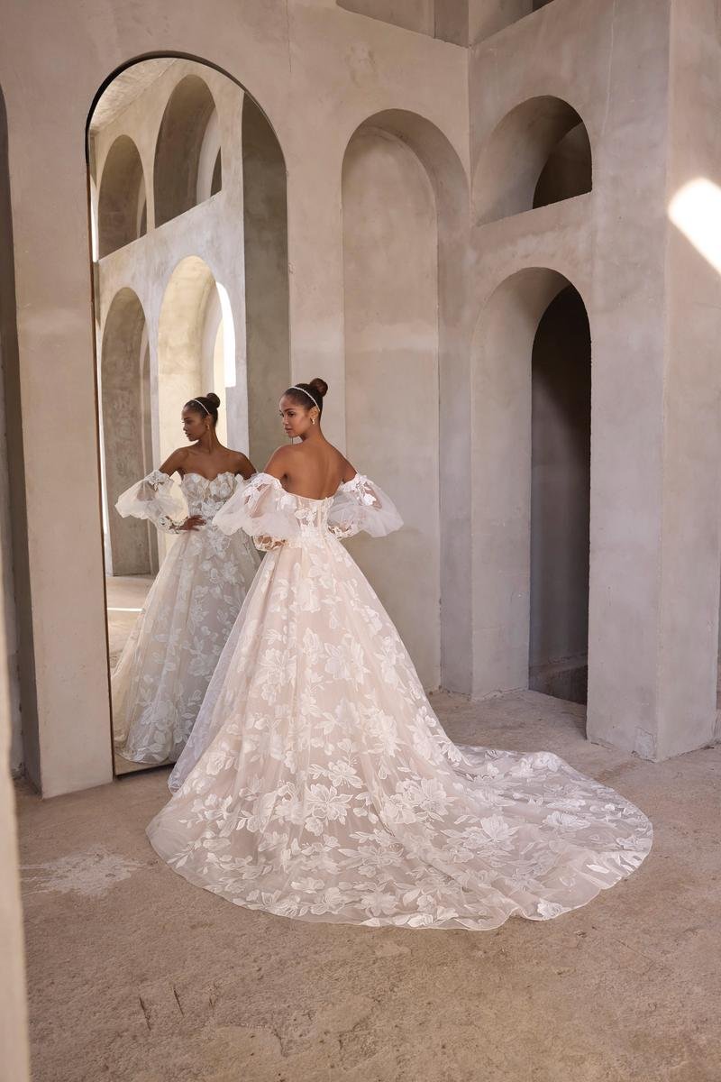 A woman in a white, floral wedding gown with off-the-shoulder puffed sleeves standing in front of a tall mirror and looking at her reflection.