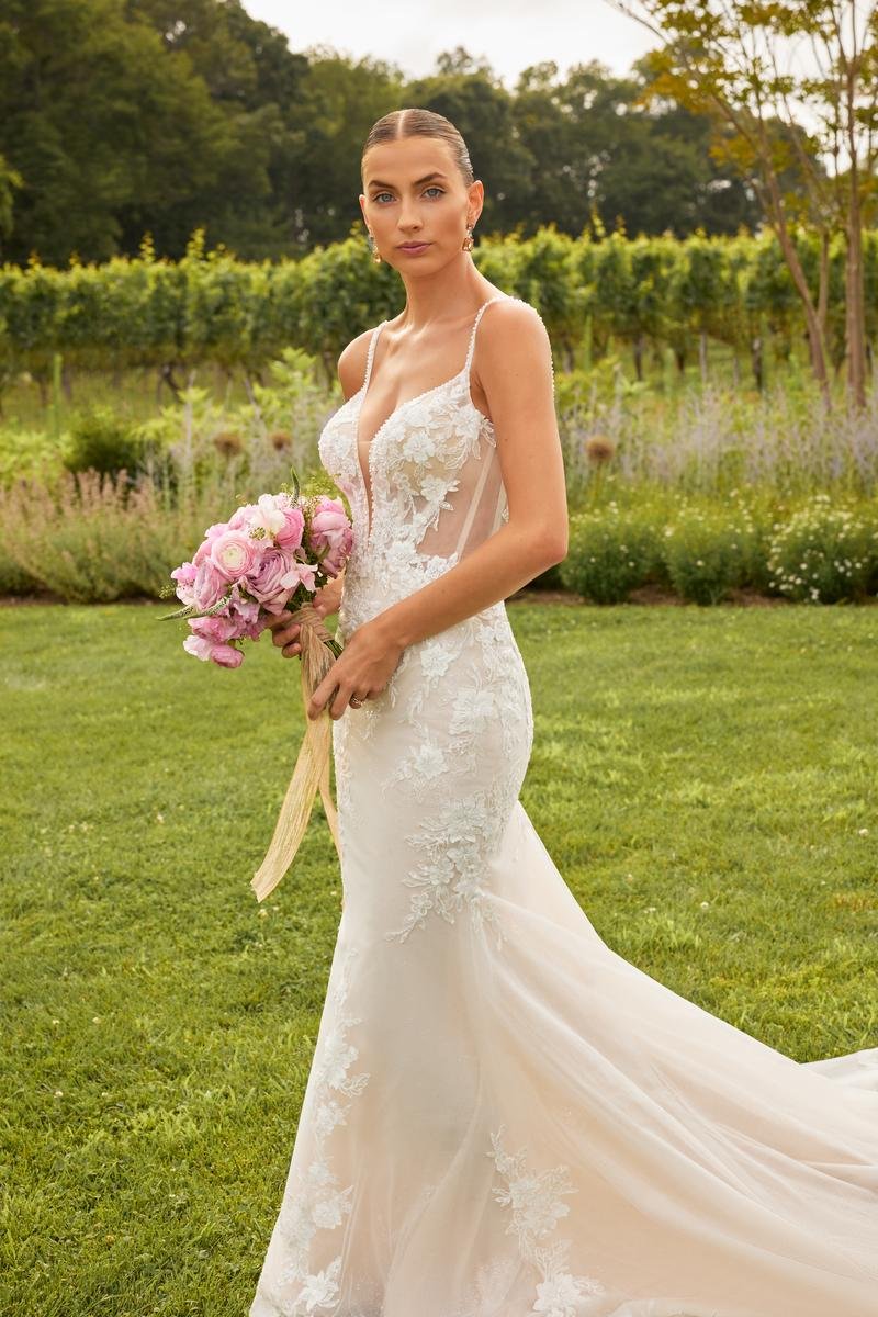 A bride in a lace wedding dress holding a pink and white bouquet standing on a grassy field with trees and bushes in the background.