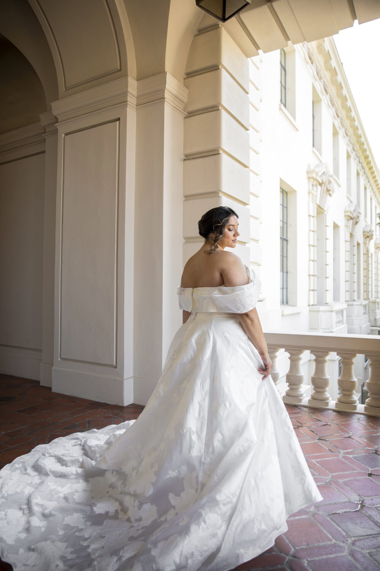 Bride in a white off-shoulder wedding gown standing on a balcony with ornate facade and large windows behind her, looking to the side.